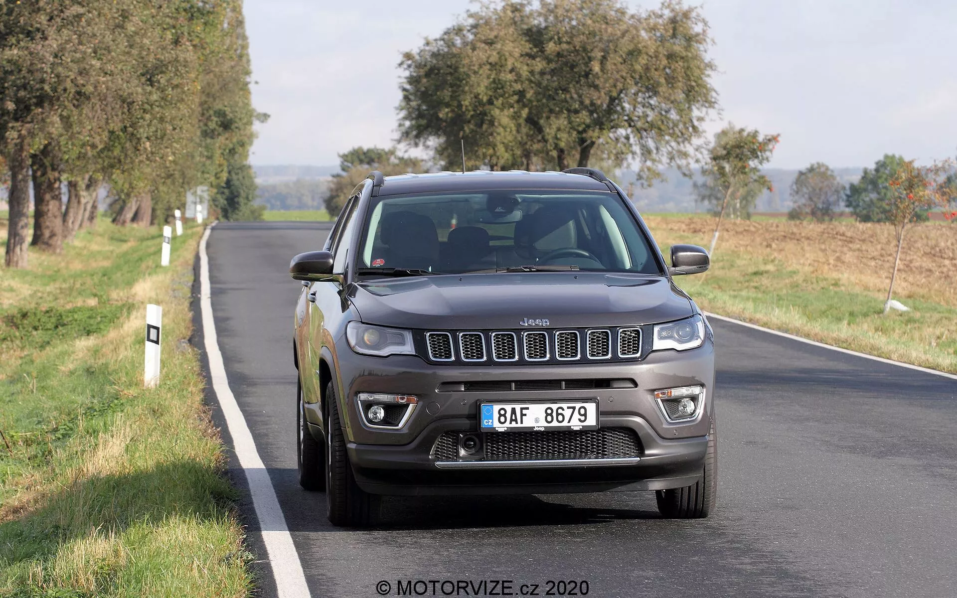 Bild av en Jeep Compass från 2017 som kör på en tvåfilig väg, fotograferad från en främre trekvartsvinkel. Fordonets ikoniska sju-slotsgaller, detaljerade strålkastare med LED-dagljusljus, dimljus och flerspakiga legeringshjul är klart synliga. Jeepens mörkgrå eller svarta färg ger den ett solitt och robust utseende, med naturligt dagsljus som lyfter fram dess funktioner. Fordonet verkar vara i rörelse, med lätt suddighet på hjulen. Träd och öppna fält i bakgrunden antyder en lantlig eller semi-lantlig miljö. Bilen upptar den centrala till nedre halvan av ramen, något till höger, och visar dess främre galler, strålkastare, stötfångare, motorhuv, förarsidans skärm, hjul och framdörr.