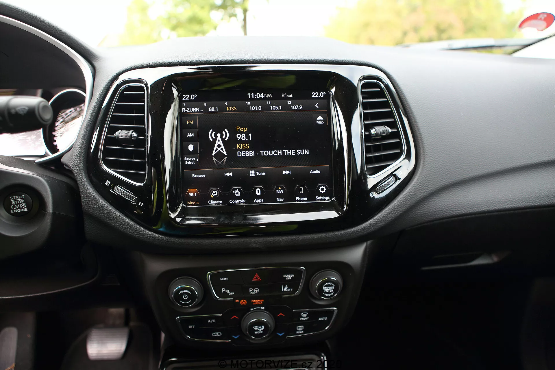 Interiören av en 2017 Jeep Compass med fokus på det centrala instrumentbrädeområdet med en stor, centralt monterad infotainment pekskärm som visar anslutningsfunktioner såsom radio, media och satellitnavigering. Under pekskärmen finns klimatsystemkontroller inklusive rattar och knappar med kromdetaljer och en LCD-display. De delvis synliga rattrarna på ratten inkluderar alternativ för farthållare och justering av ljudvolym. Ytterligare designfunktioner som visas inkluderar vertikala ventiler, sätesvärmekontroller och en USB-port, som framhäver fordonets interiörteknik och estetiska design.