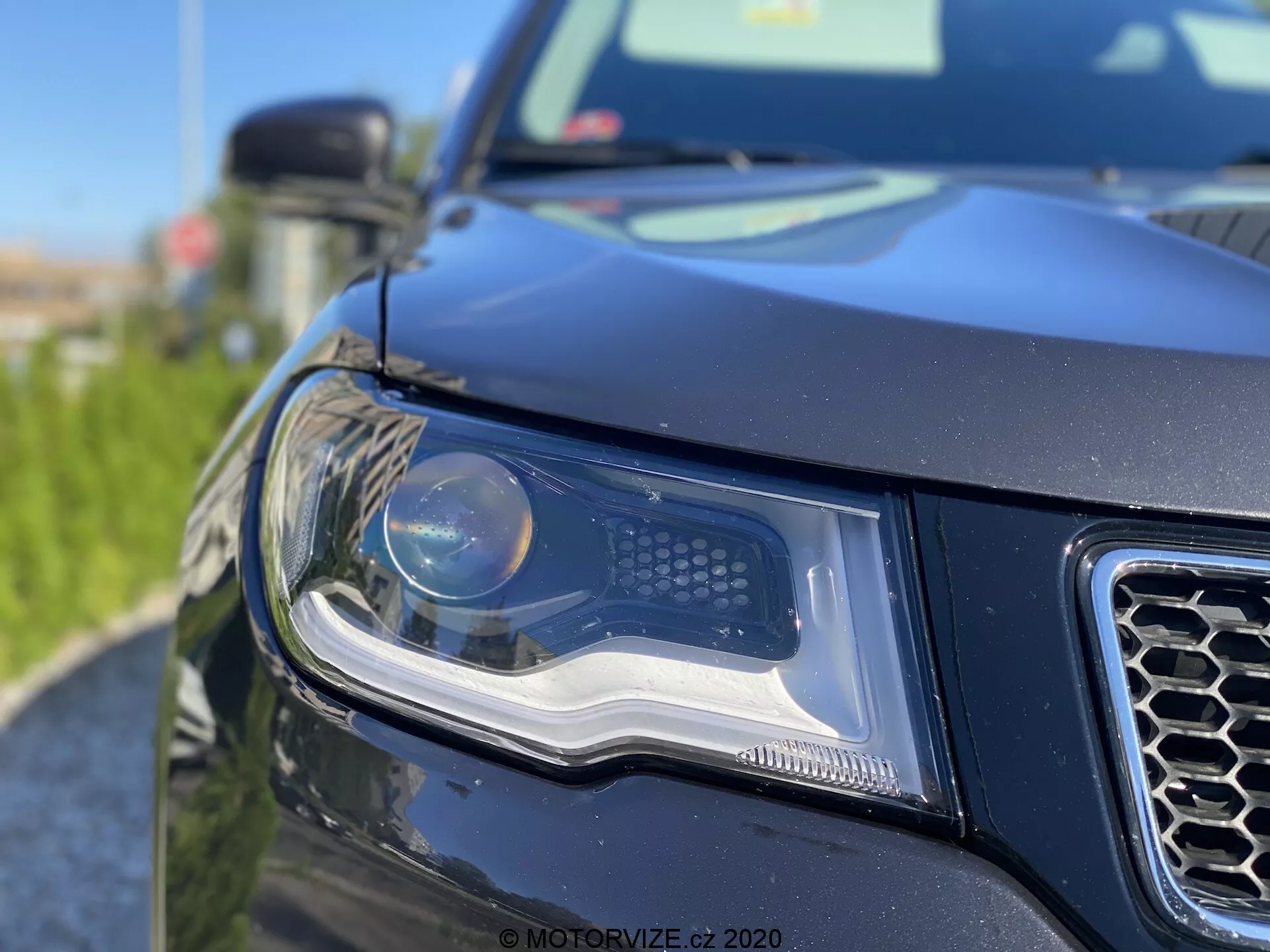 Närbild på vänster framhörn av en modern SUV, med fokus på avancerad strålkastare med projektor- och LED-dagsljus. Bilden visar del av honungskakemönstrad grill, svart eller mörkfärgad kaross med kraftig, konturerad design och delvis vy av stor sidospegel i dagsljus med goda ljusförhållanden. Oskarp vegetation i förgrunden tillför djup och en konstnärlig touch.