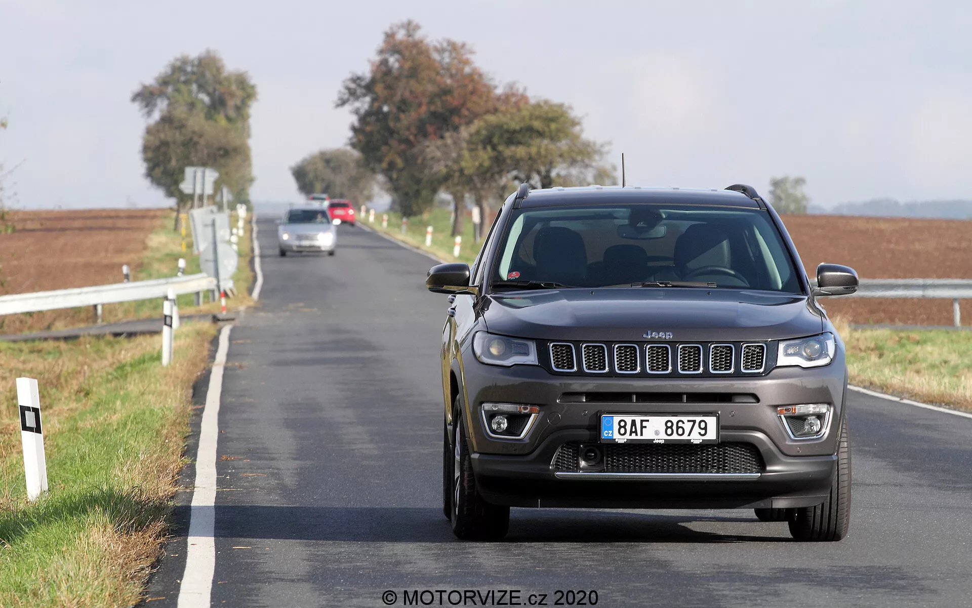 Låg vinkelbild av en Jeep Compass 2017 som kör mot kameran på en tvåfilig asfalterad väg. Frontalvyn visar grillen, strålkastarna och stötfångaren i detalj med synliga moderna funktioner som LED- eller HID-lampor och främre parkeringssensorer. Bilen är i rörelse med en suddig landlig bakgrund som har vita vägposter och öppna fält under en klar himmel.