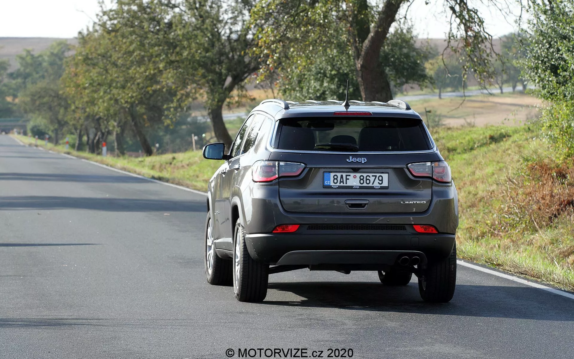 Bakifrånvy av en Jeep Compass SUV från 2017 i metallicgrå färg, som kör bort på en tvåfilig väg, placerad något till höger i ramen. Fordonet, i höger körfält, har synliga funktioner som baklucka med Jeep-märke och registreringsskylt, bakruta med en liten spoiler, baklyktor med röda och vita sektioner, bakre stötfångare med sensorer och en hajfensantenn på taket. Ljushjulen är delvis synliga. Scenen antyder en solig dag med omgivande träd och gräs, vilket tyder på en lantlig eller halv-lantlig miljö.