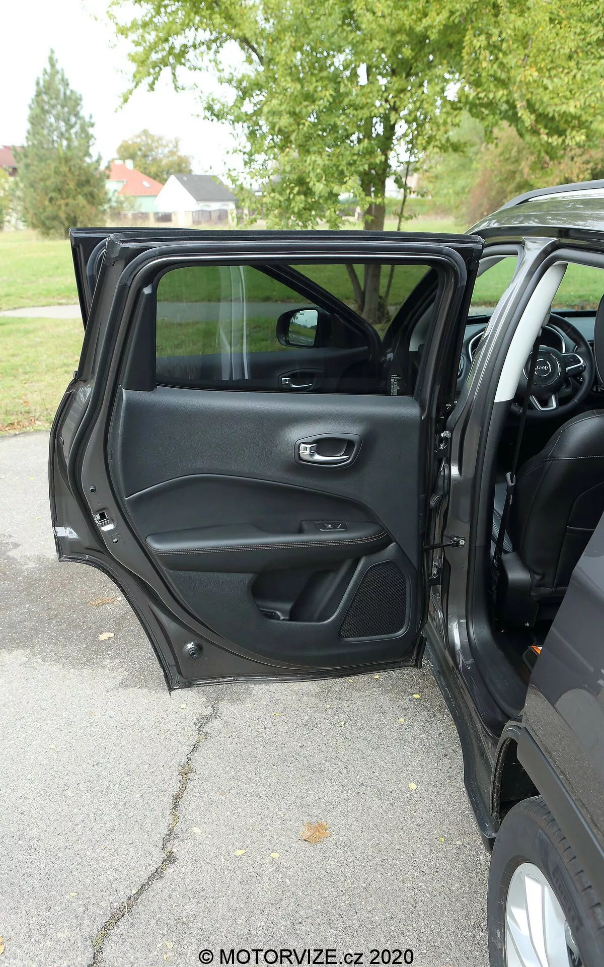 Interiör vy av en Jeep Compass 2017 från utsidan som visar den öppna förardörren med ett metalliskt och ergonomiskt handtag, förvaringsfack, högtalargaller och kontrollpanel för fönster- och dörrlåsoperationer. En del av instrumentbrädan, mittkonsolen och den multifunktionella ratten är synliga, vilket antyder en modern och funktionsrik interiör. Naturligt ljus, mulet väder, träd i bakgrunden vilket tyder på en utomhusmiljö i en park eller trädklädd gata.