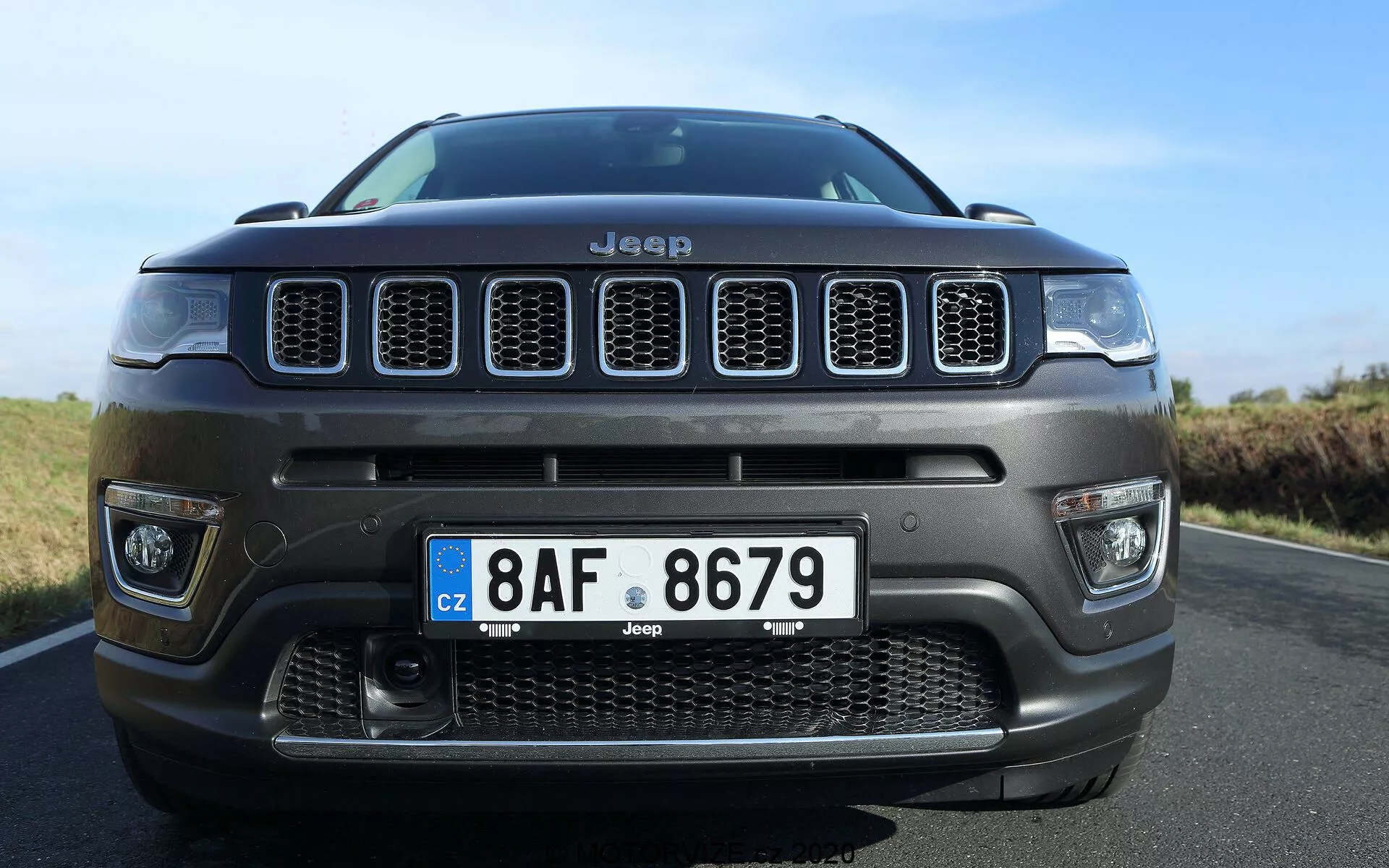Låg vinkel framifrån av en Jeep Compass 2017, med betoning på den karakteristiska sjuslotsgrillen och centrala främre stötfångaren med ett stort luftintagsgaller. Närbildsfotot, något förskjutet åt höger, framhäver fordonets imponerande hållning med fokus på grillen, detaljer av stötfångaren, europeisk stilregistreringsskylt och robusta designinslag, vilket tydligt visar Jeepens identitet och terrängförmåga.