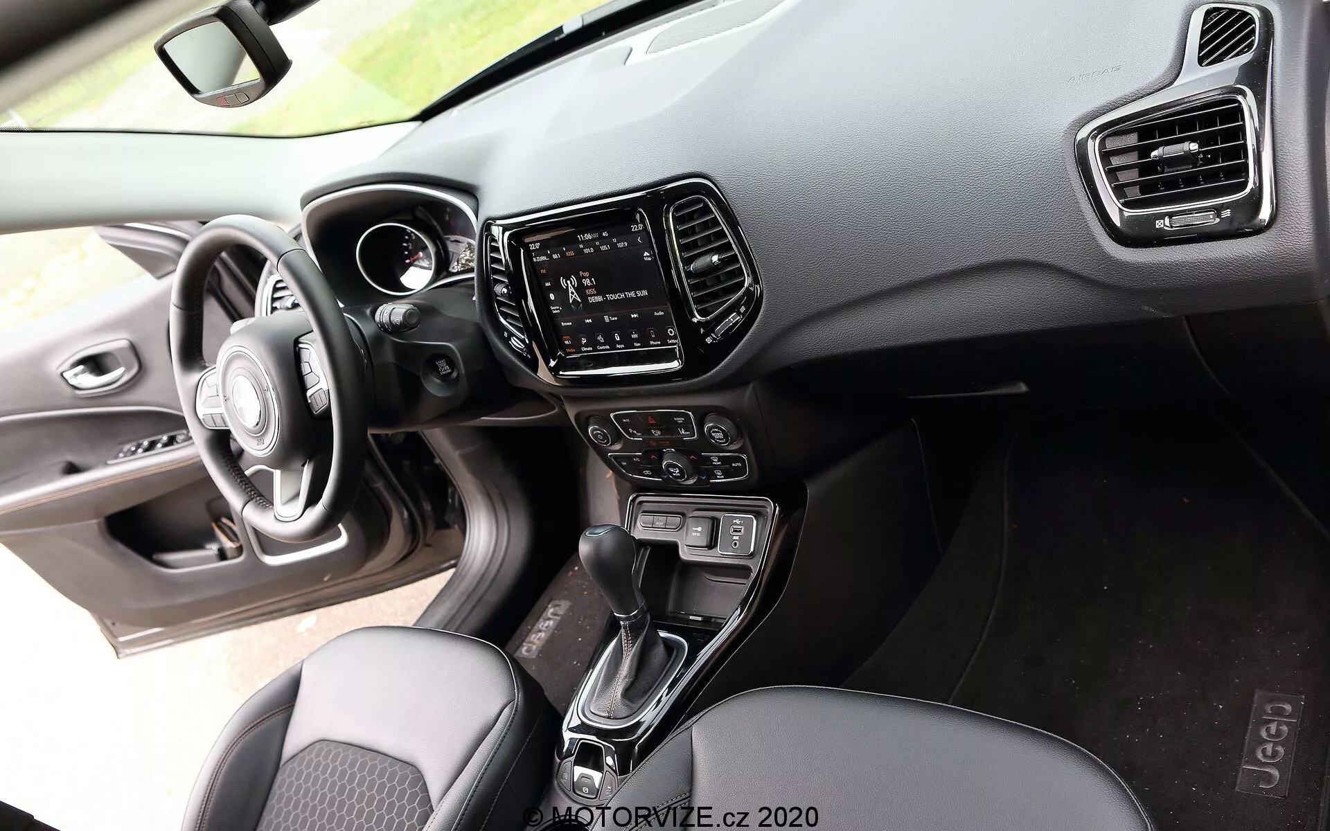 Interiör vy av en Jeep Compass 2017 från passagerarens perspektiv, som visar en tre-ekrad ratt med integrerade kontrollknappar, en delvis synlig digital instrumentpanel på instrumentbrädan gjord av mjuka material och hård plast med synliga sömmar, ett infotainmentsystem med pekskärm i mittkonsolen flankerat av vertikala klimatanläggningsventiler, en automatisk växelspak mellan framsätena, knappar och rattar för klimatanläggning, tygsäten med ett mönstrat design och en glimt av förardörrens panel med justeringskontroller för fönster och sidospeglar förstärkta av omgivande belysning, som belyser fokus på komfort och teknik i marknadssegmentet för mellanklass till högklass.