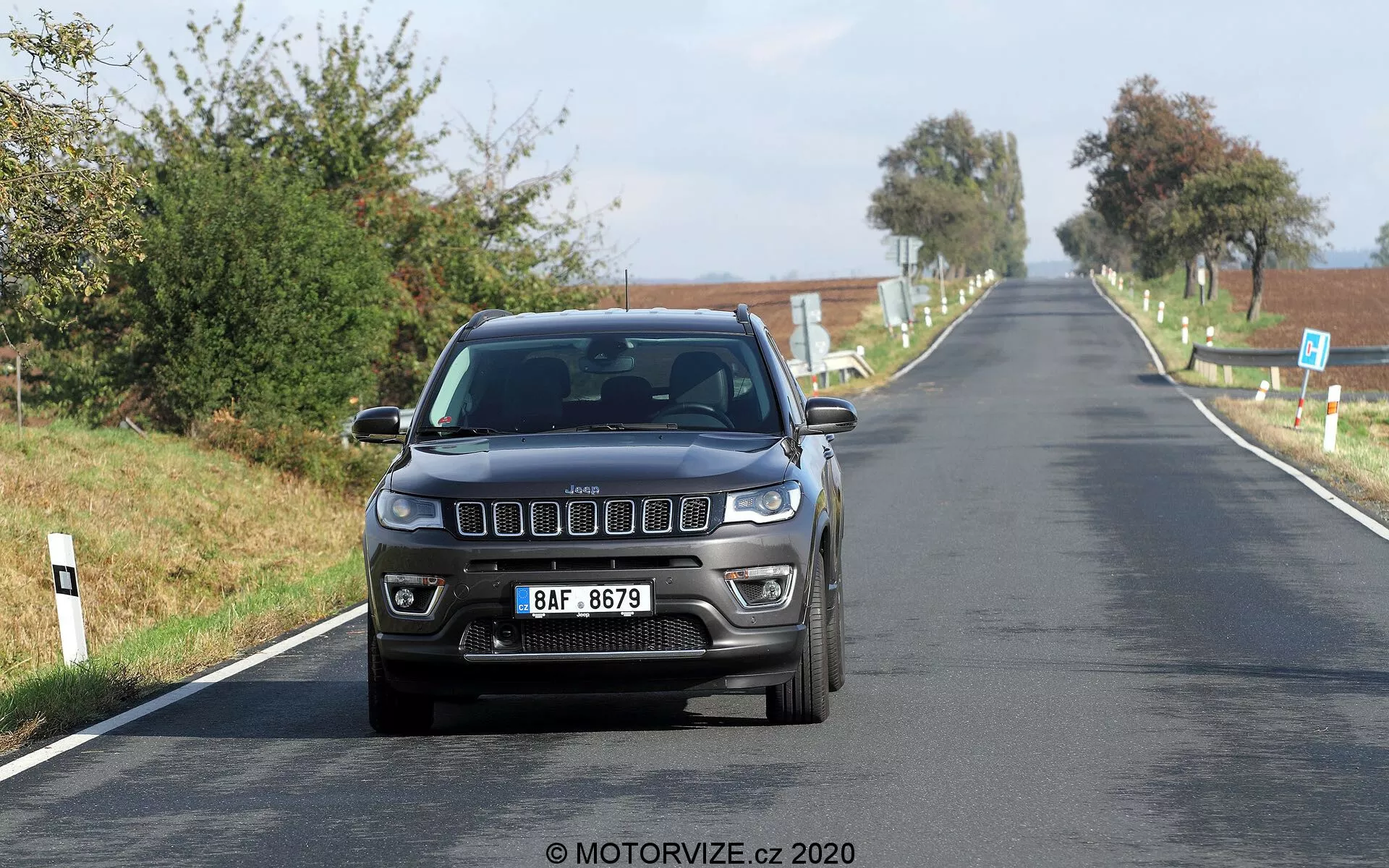Frontal vy av en Jeep Compass 2017 som kör i moderat hastighet på en landsväg. Bilden fångar de distinkta främre dragen inklusive den ikoniska sju-slots grillen, moderna rektangulära strålkastare samt dimljus integrerade i stötfångaren. Bilen är fotograferad i ögonhöjd vilket framhäver dess exteriördesign och belysningsteknik under bra naturligt dagsljus. En europeisk registreringsskylt är centralt monterad framtill, med tydlig landsbygdsvy i bakgrunden.