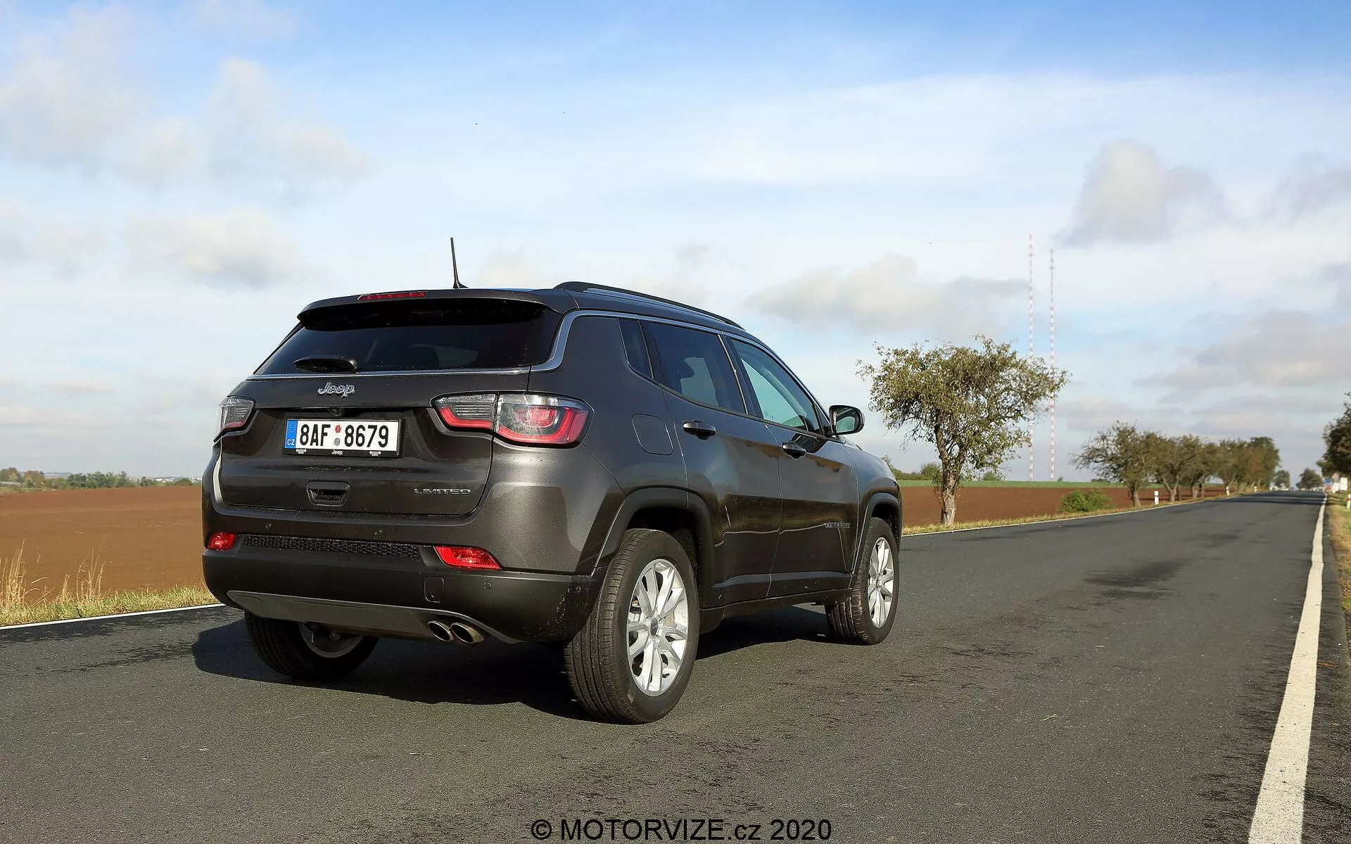 En bakre fyra kvarts vy av en 2017 Jeep Compass. Bilden framhäver bilens sidprofil och bakdel, med framträdande baklyktor, en bakre vindrutetorkare, en takspoiler, reflexer på den bakre stötfångaren och flerspokedesignade aluminiumsfälgar. Fordonet är parkerat vid vägkanten bredvid en gräsbevuxen kant. Himlen är delvis molnig, vilket tyder på varierande väderförhållanden.
