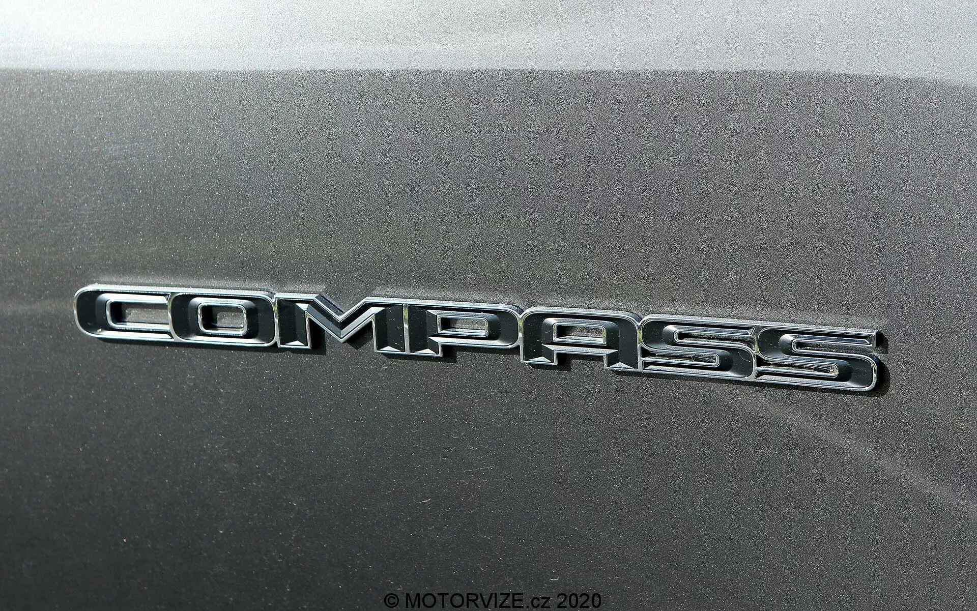 Närbild av Jeep Compass 2017-märket med glänsande, kromad inskription mot strukturerad bilpanel.
