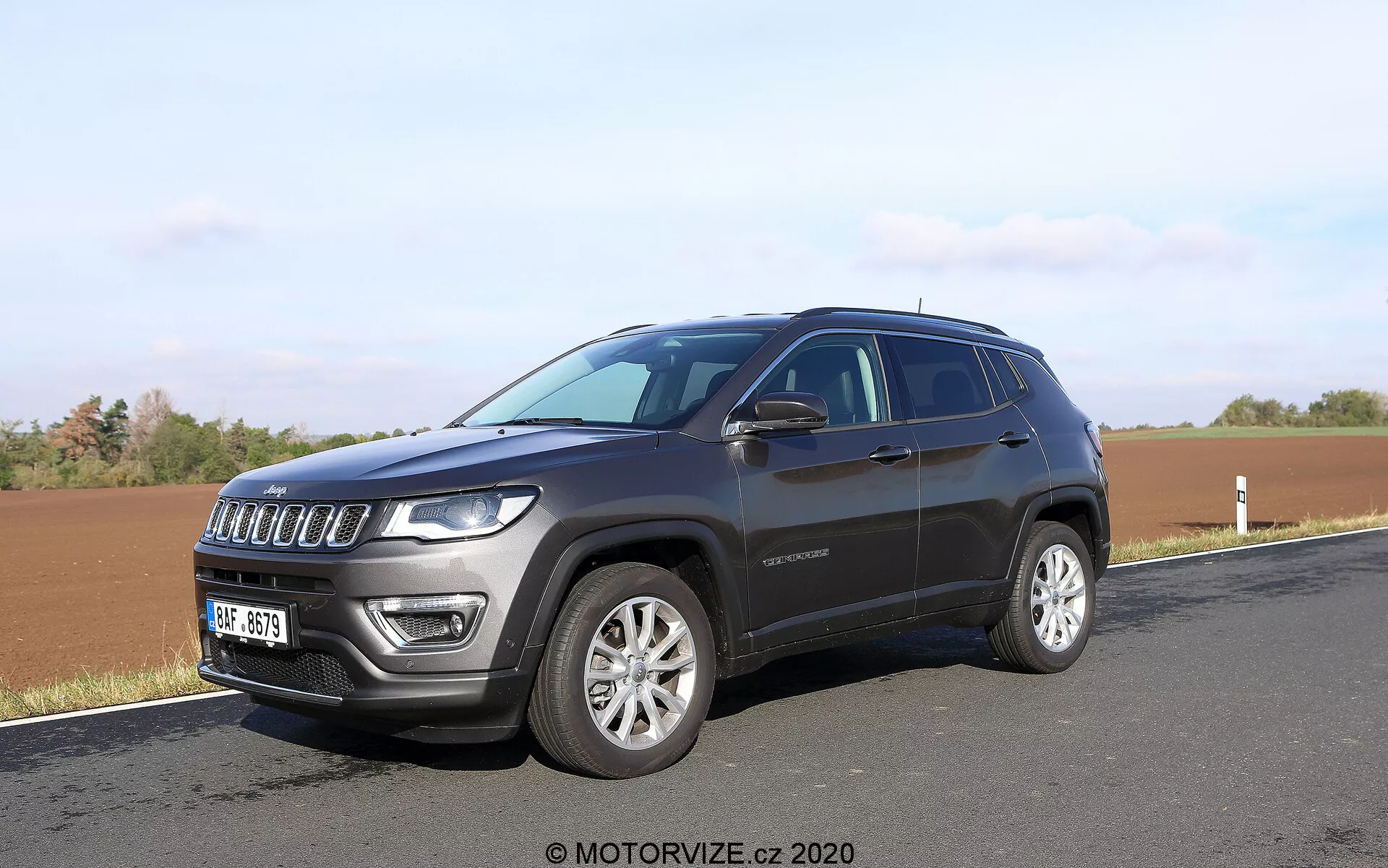 Modern SUV parkerad vid en väg med en åker i bakgrunden, fotograferad från en vinkel på ungefär 45 grader. Bilden framhäver SUV:ns sju-slotsgrill, distinkta LED-utrustade strålkastare, legeringsfälgar och mörk metalliclack. Sidoelement som fönsterrad, dörrhandtag, troligen integrerade blinkers i sidospeglarna och en något sluttande taklinje är synliga. Ett emblem på framdörren är också synligt, vilket kan ge information om modell eller utrustningsnivå.