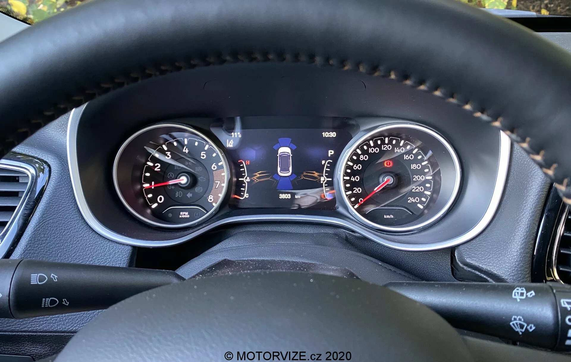 Närbild av Jeep Compass 2017:s instrumentpanel från förarens perspektiv, som visar både analoga mätare och en central digital display. Funktioner inkluderar en varvräknare, hastighetsmätare med dubbla enheter, växelvalgsindikator, bränslemätare, vägmätare och ytterligare säkerhetsteknikindikatorer som filhållningsassistans. Sömmen på ratten i läderliknande material betonar kvalitetsmaterial och komfort, medan styrspakar och knappar antyder avancerade bekvämlighets- och hjälpsystem för förare.