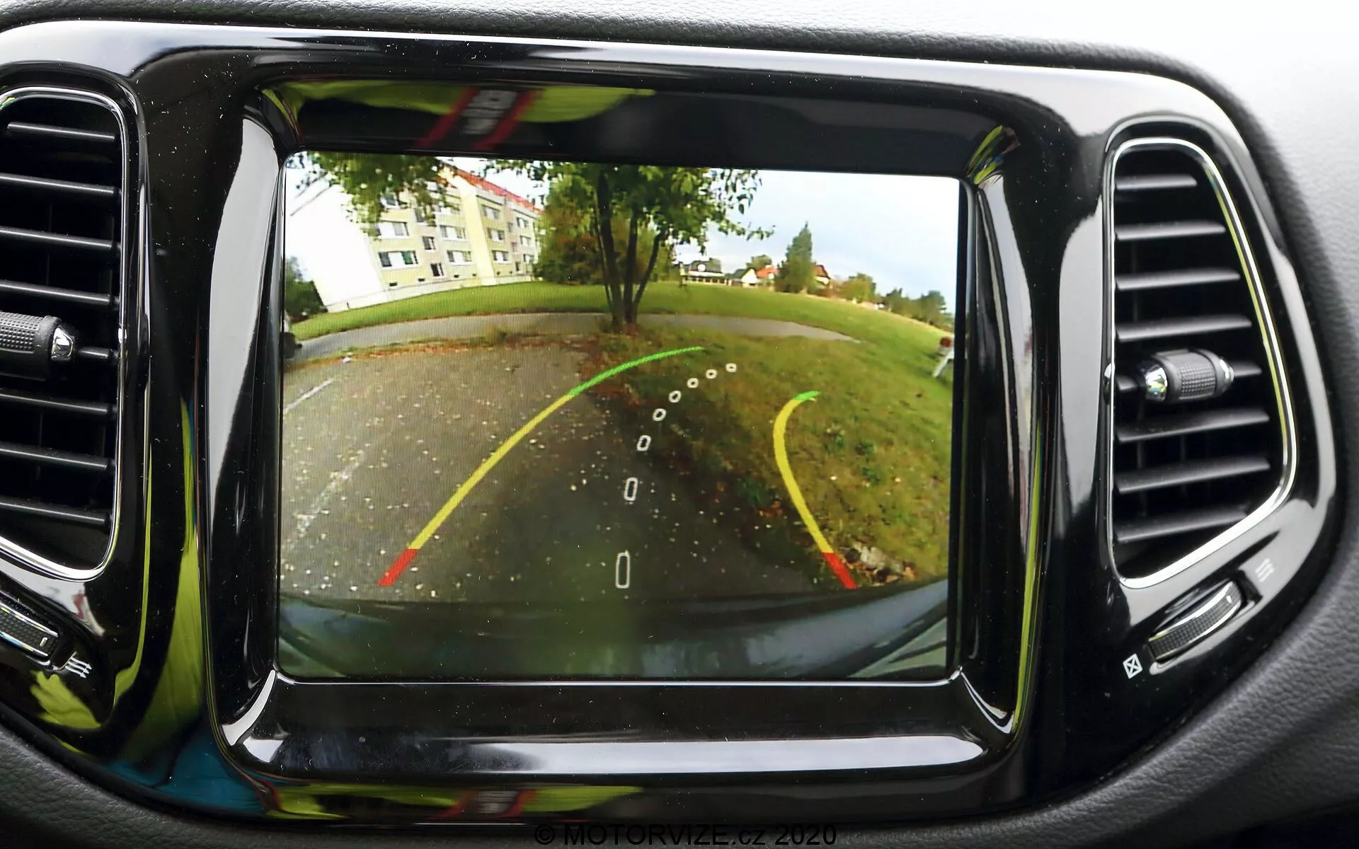 Bild av ett backkamerasystem på instrumentpanelens skärm i en Jeep Compass 2017. Kameran, placerad bak vid registreringsskylten, ger en vidvinkelbild bakom fordonet. Synliga är överlagda riktlinjer för backning, som anger fordonets bredd och förväntad bana med gula och röda linjer för olika avstånd. Scenen visar en parkeringsplats eller uppfart med träd och gräsmatta i bakgrunden. Skärmen är centralt placerad på instrumentpanelen, omgiven av luftkonditioneringsventiler, och har en tydlig bildkvalitet, typisk för modern fordonsteknik.
