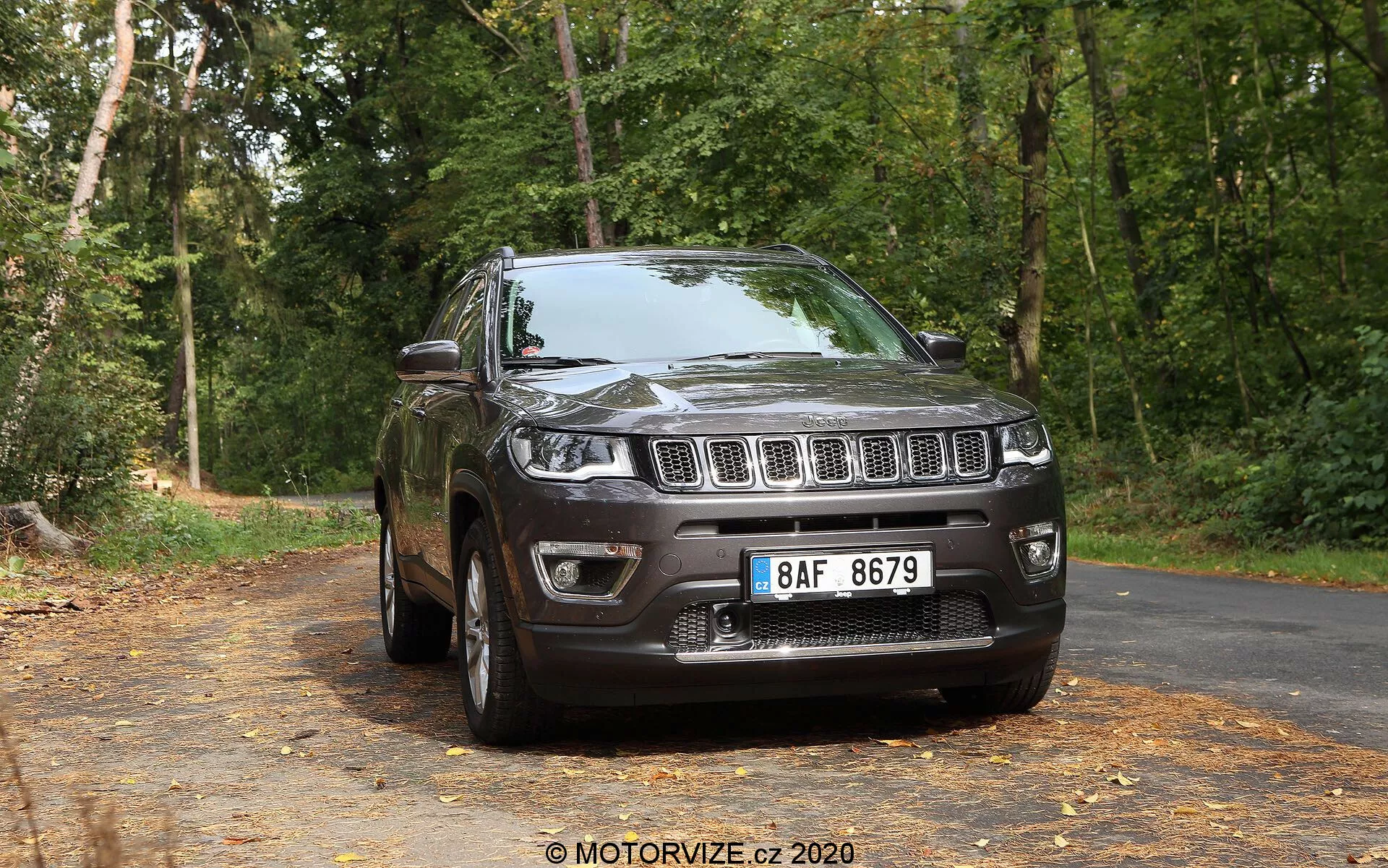 Trefjärdedels vy framifrån av en mörkfärgad Jeep Compass SUV från 2017 på en tyst, lövtäckt asfalterad väg med ett bakgrund av träd i en skogsmiljö. Bilden visar den främre grillen med distinkta vertikala spår, flankerad av strålkastare som sträcker sig in i skärmarna, en väl definierad främre stötfångare med dimljus och en formad förhöjd motorhuv. Sidoprofilen visar lättmetallfälgar, en synlig skärm, sidospeglar, dörrhandtag, fönsterkonturer och ett takräckessystem, allt belyst av naturligt ljus.