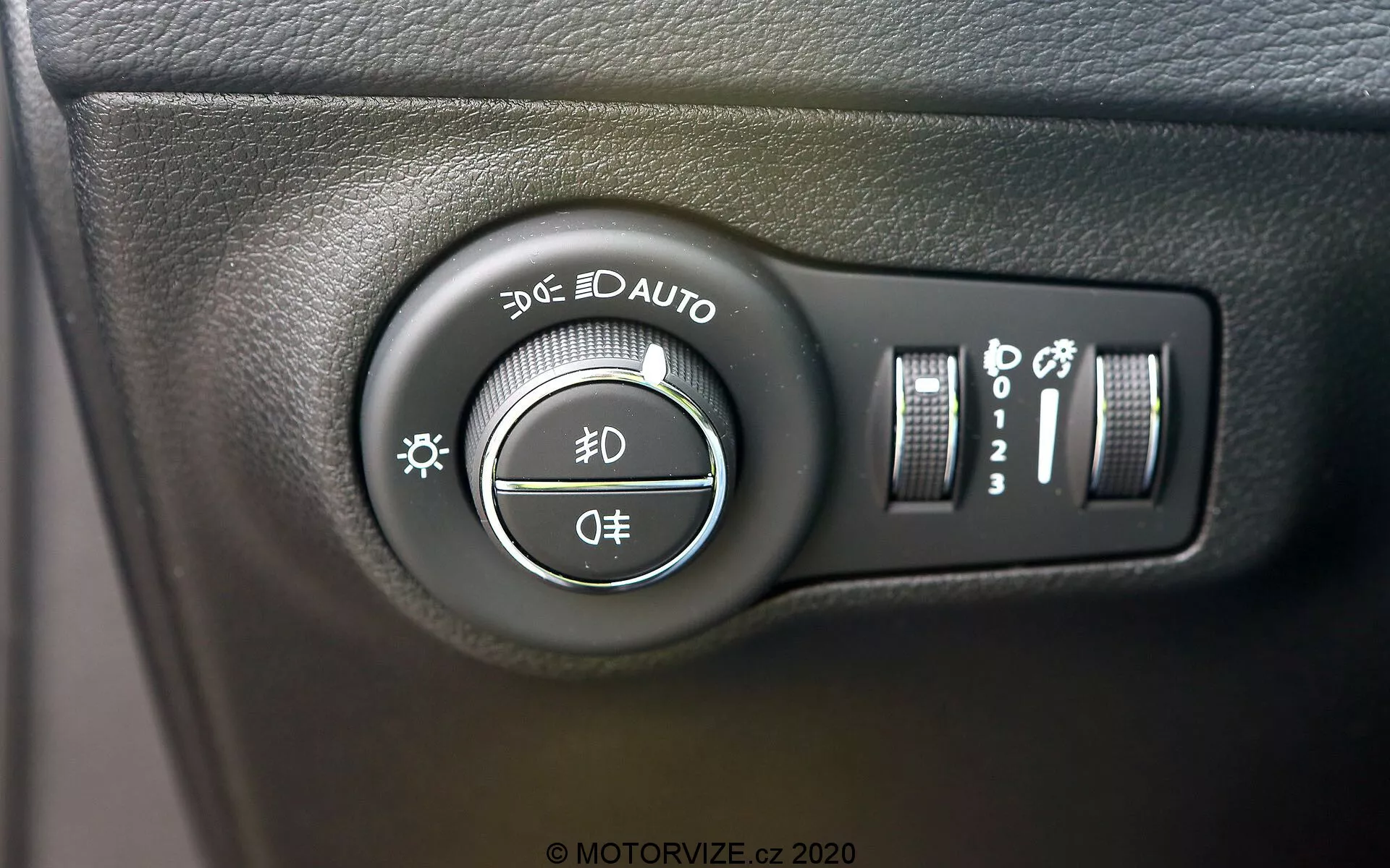 Närbild på strålkastarkontrollen på Jeep Compass 2017, placerad på instrumentbrädan till vänster om ratten. Bilden, tagen i en lätt vinkel, betonar vridreglaget med inställningar för parkeringsljus, dagljus, automatiska strålkastare, halv- och helljus samt en avstängningsposition. Ytterligare kontroller till höger inkluderar en vertikal skjutreglage för instrumentbrädans ljusstyrka och en knapp som kan indikera automatiska helljus eller strålkastarjusteringssystem. Strålkastarkontrollen sitter på en texturerad svart plastyta med tydliga vita märken och lätt blanka reflektioner.