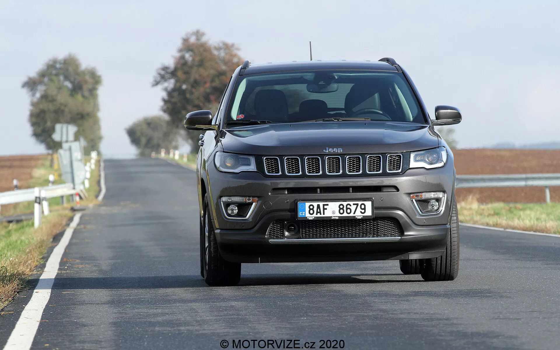 Frontal vy av en rörlig 2017 Jeep Compass på en tvåfilig väg, fotograferad från ett lågt perspektiv med fokus på den framträdande sju-spårsgrillen, fyrkantiga strålkastarna med moderna dagsljuslampor och synliga dimljus och främre parkeringssensorer på stötfångaren. Vägen sträcker sig mot horisonten med en solid dubbel gul linje som indikerar en förbjuden överkörningszon. Den oskarpa bakgrunden antyder att fordonet är i rörelse och visar dess förhöjda hållning som är typisk för SUV:ar.