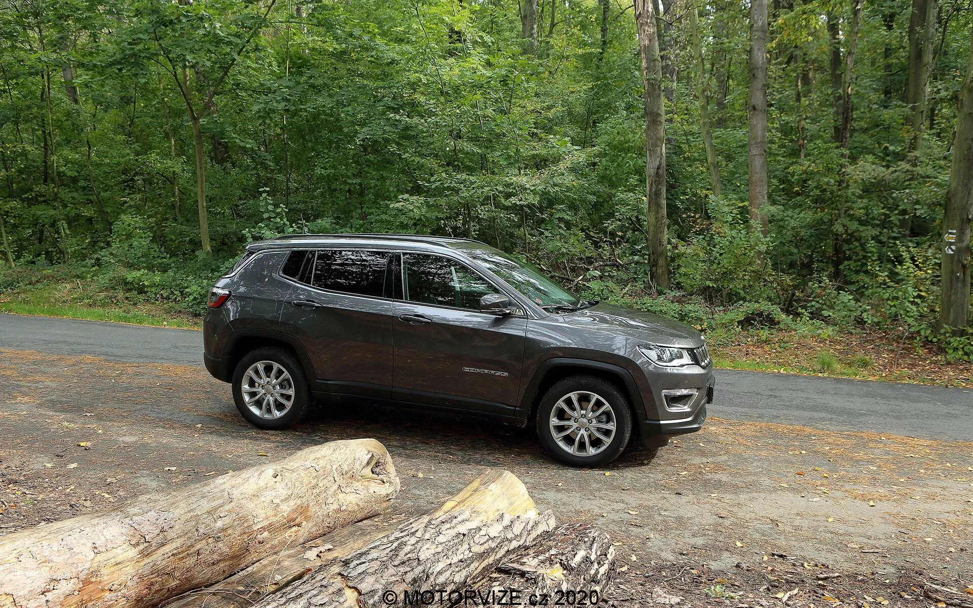 En sidovy av en Jeep Compass 2017 SUV parkerad på en asfalterad väg i en naturlig miljö med frodiga gröna träd. Fordonet visar moderna designlinjer längs dörrarna och hjulhusen, med synliga flerspokedesignade fälgar. Den har hög markfrigång som antyder terrängegenskaper. Bilden fångar en del av frontgrillen och strålkastarna, vilket tyder på en modern stil. Takräcken, integrerade blinkers i sidospeglarna och tonade fönster är närvarande. Den mörk färgen med en blank yta antyder ett väl underhållet fordon. Den stående SUV visas i riklig naturbelysning, perfekt för att visa dess egenskaper och lockelse för utomhus- och äventyrsintresserade.