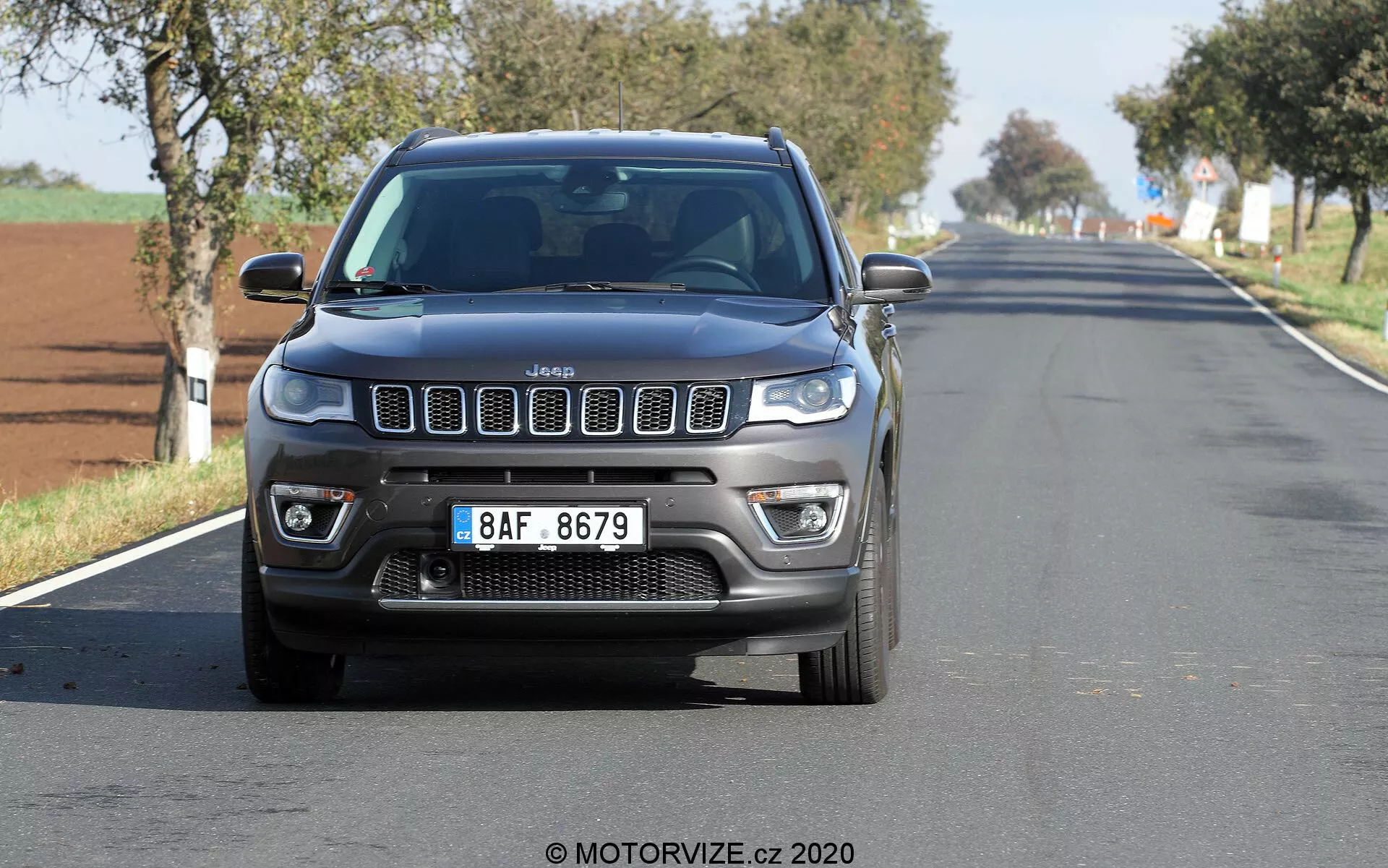 Frontal vy av en Jeep Compass 2017 som färdas på en tvåfilig landsväg eller förortsväg, fångad från en låg vinkel, som visar dess ikoniska sju-slot-galler, LED-dagsljuslampor, dimljus och robusta motorhuvkonturer. Bilen är centrerad och relativt nära kameran, med en lätt rörelseoskärpa på hjulen och bakgrunden. Klara himlar antyder en ljus, solig dag.