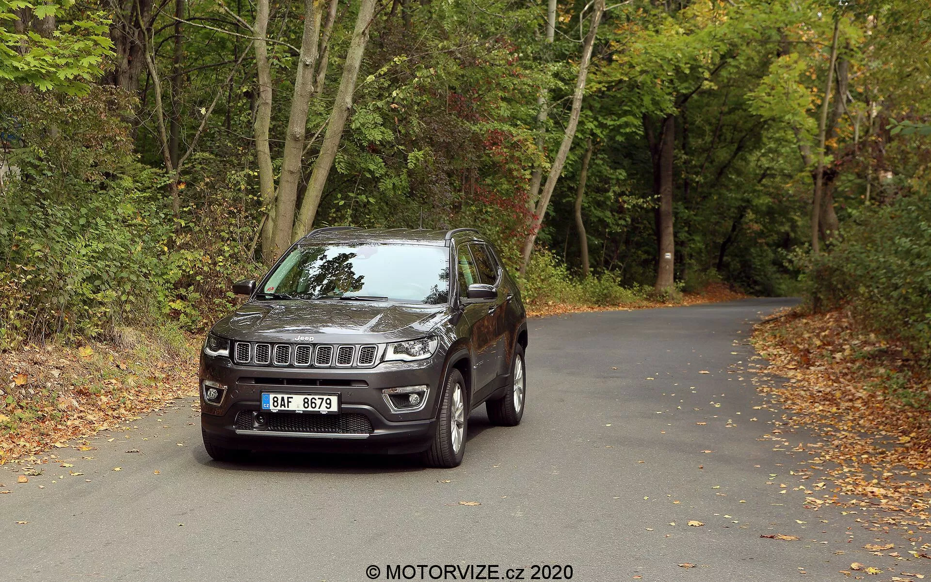 En 2017 Jeep Compass SUV placerad på en asfalterad väg omgiven av skogar med löv på marken, vilket antyder höst. Fordonet är fotograferat från en främre tredjedel vinkel, med den distinkta Jeep sjuslotsgrillen och mittensymbolen, moderna LED-strålkastare och dimljus integrerade i stötfångaren. Det har lättmetallfälgar, sidospeglar med inbyggda blinkers, robust konstruktion med hjulbågsplaster och lätt tonade fönster. Den detaljerade utsikten framhäver design- och teknoelement som antyder dess lämplighet för både stads- och terrängäventyr.