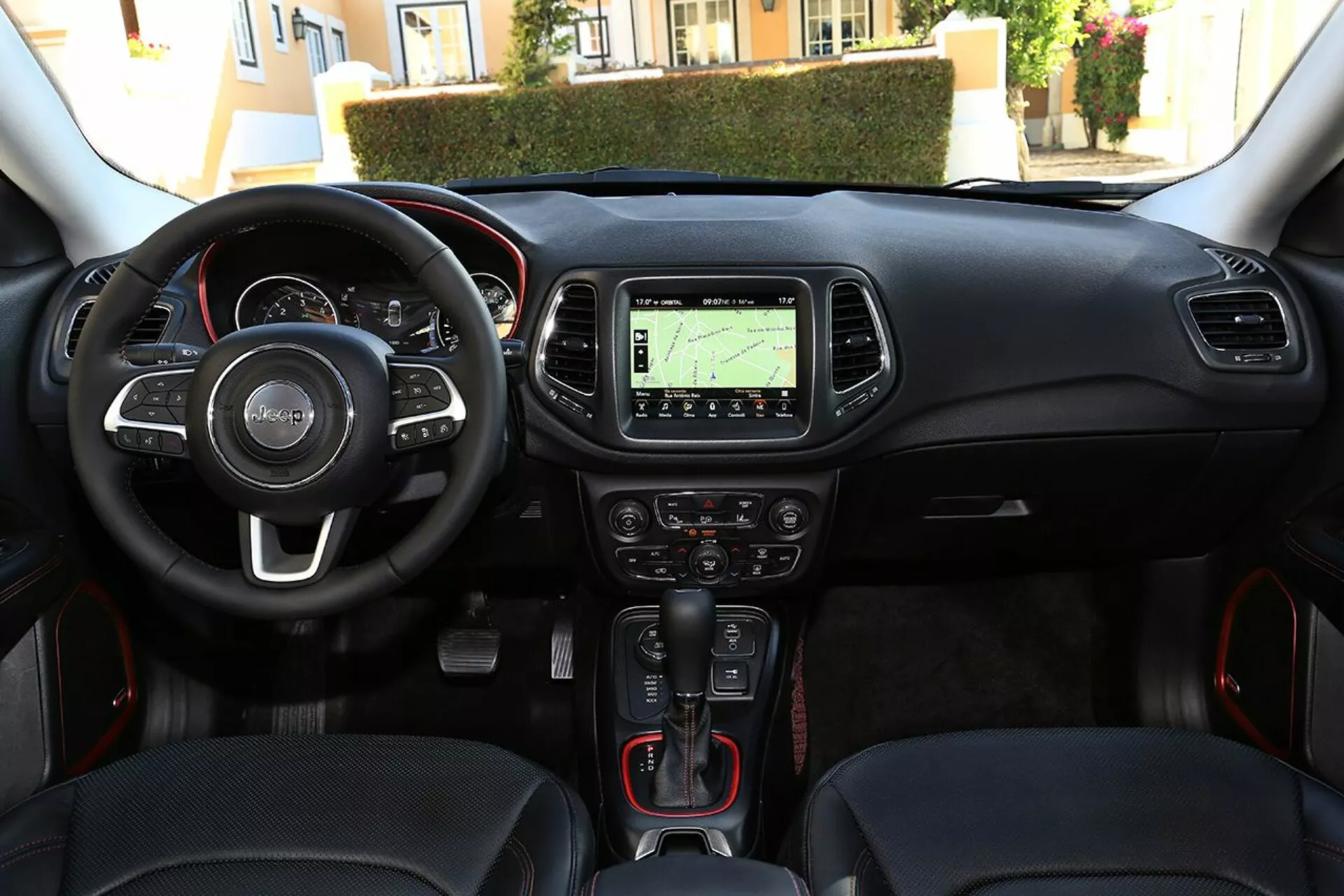 Interiörvy av Jeep Compass 2017 från förarens perspektiv som visar ratten med monterade kontroller, instrumentkluster, mittkonsol med navigeringssystem, klimatkontroller, automatisk växelspak med sportiga röda detaljer, nyckellös start-stopp-knapp och multimediaanslutningsport