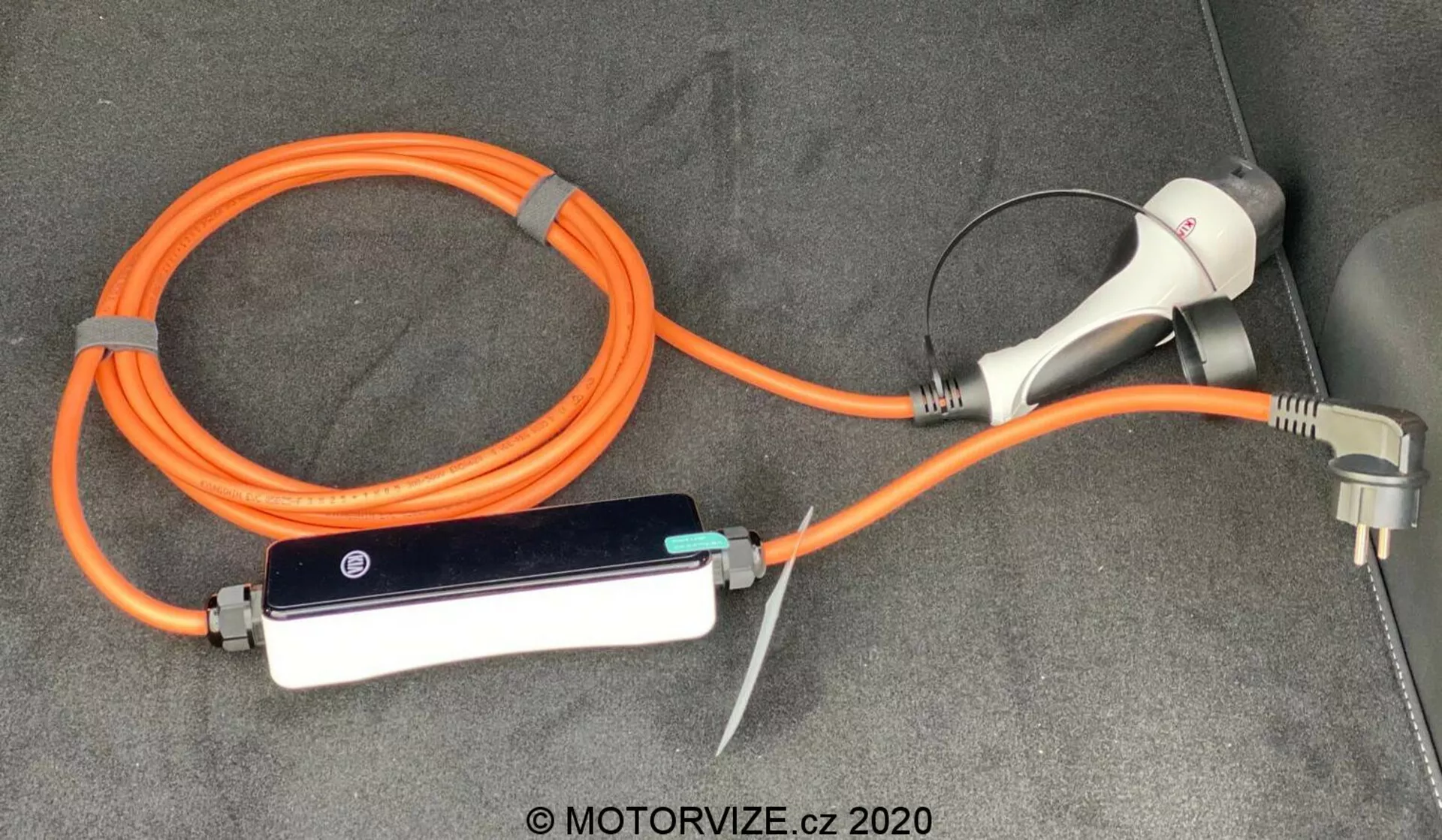 Elektrisk fordon (EV) laddkabel och adapteruppsättning för Kia Ceed 2018, med en portabel orange EVSE (Elektriskt Fordonsförsörjningsutrustning) med hög synlighet för säkerhet. Uppsättningen innehåller en laddningskontakt och stickpropp för hemmabruk, samt en inline-kontrollbox med lysdioder, allt utlagt på den matta golvytan i ett fordons bagageutrymme eller lastutrymme.