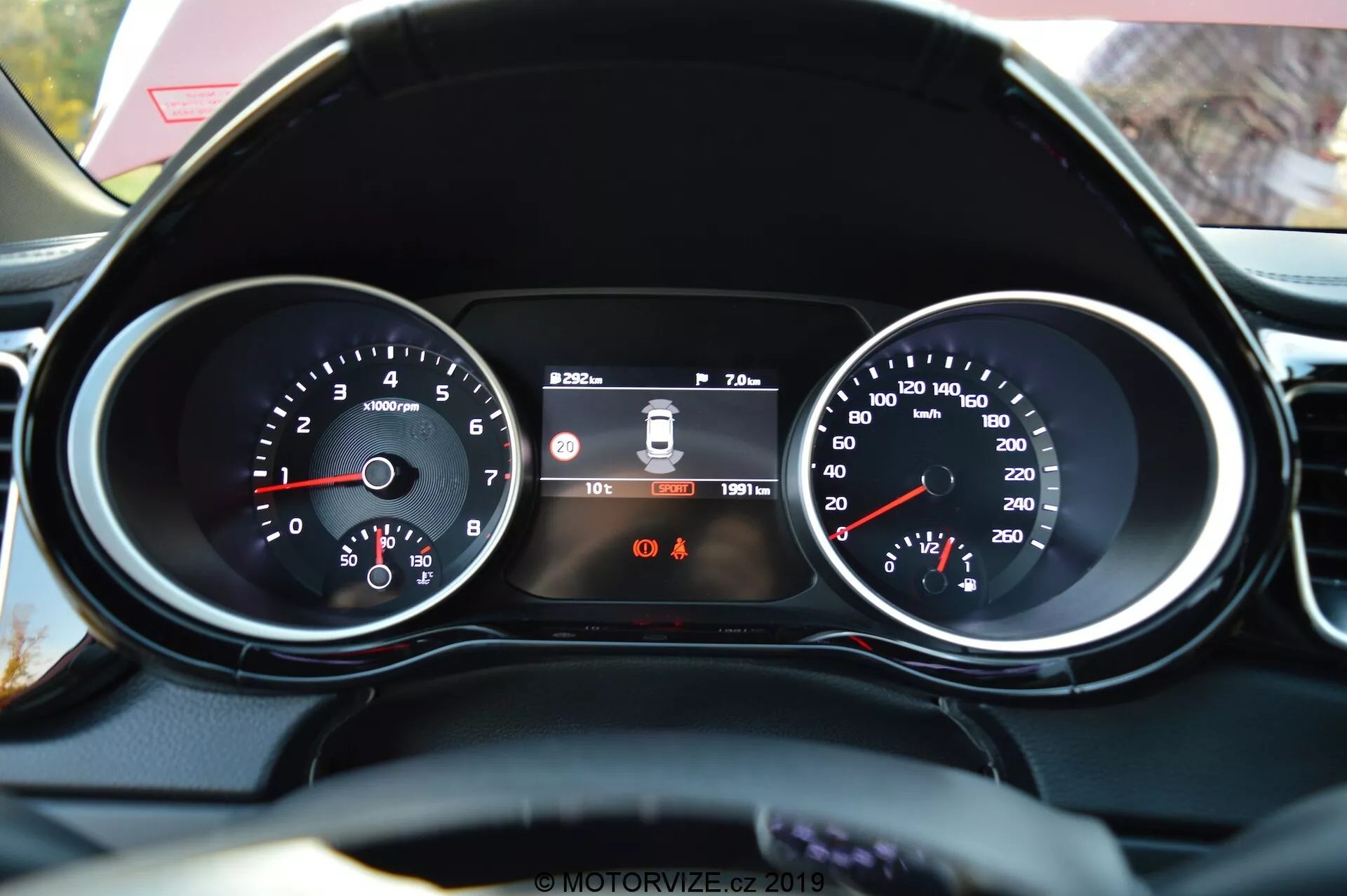 Närbild från förarens perspektiv av instrumentpanelen och en del av den läderklädda ratten i Kia Ceed 2021. Funktioner inkluderar en vänster varvräknare med RPM-avläsning, en central digital display som visar körfältsavvikelser, tid och vägmätare, och en höger hastighetsmätare med hastighet i km/h. Under den centrala displayen syns två varningsindikatorlampor, där säkerhetsbältespåminnelsen är identifierbar.