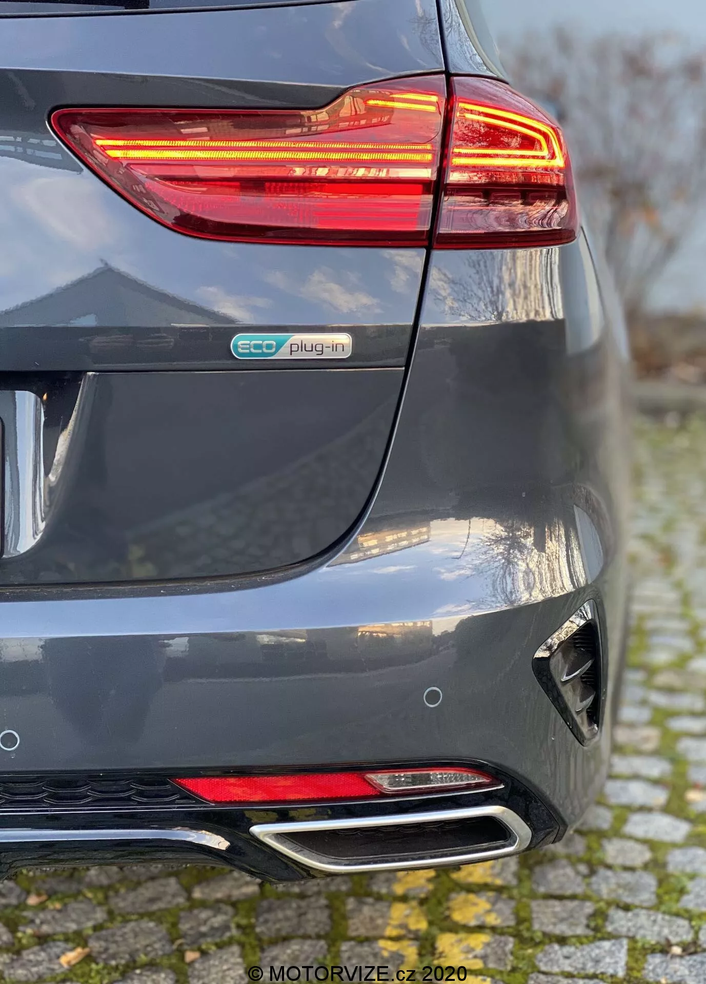 Detaljerad vy av den bakre delen av en 2018 Kia Ceed i metallisk grå, med fokus på det upplysta LED-bakljuset, eco plug-in-märket, skulpterad bakre stötfångare med reflex och diffusor, samt dubbla kromade avgasrör. Perspektiven visar sammanflödet av den bakre stötfångaren och högra sidan, vilket betonar hybrid-elektriska fordonselement och moderna designegenskaper.