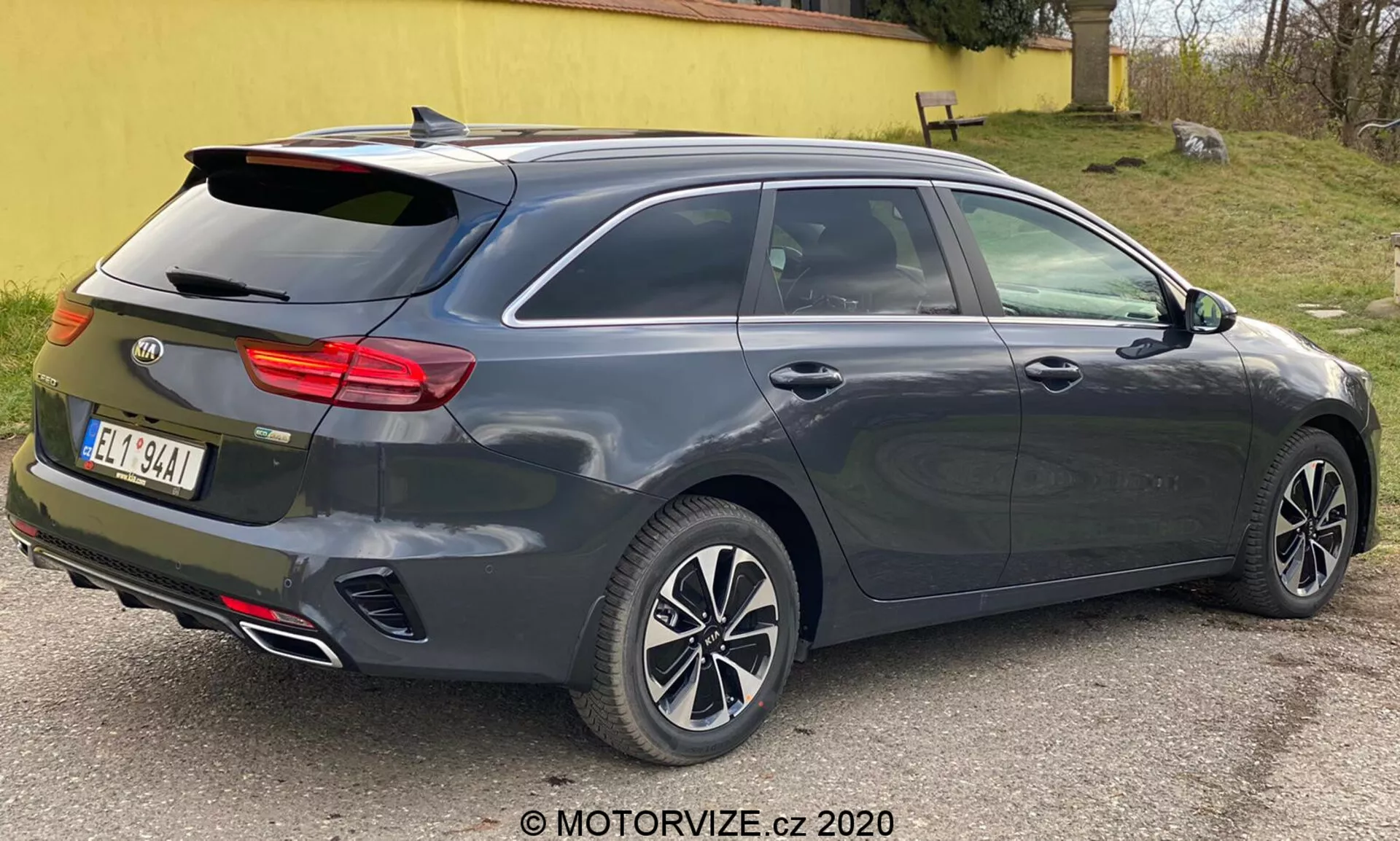 En mörkgrå metallic Kia Ceed 2018, visad från trekvart bakifrån, som framhäver dess moderna halvkombi- eller kombidesign med lutande bakruta, integrerad bakspoiler, flerspoksfälgar av lättmetall, dörrhandtag och sidospeglar i karossens färg troligen med integrerade blinkers, distinkt horisontellt baklyskluster möjligen med LED-teknik, hajfenantenn, dubbla avgasrör, högmonterat tredje bromsljus, avfrostningsfunktion på bakrutan, sensorer eller reflexer som indikerar ett parkeringshjälpsystem bak, fotograferad på nära håll med neutral belysning.
