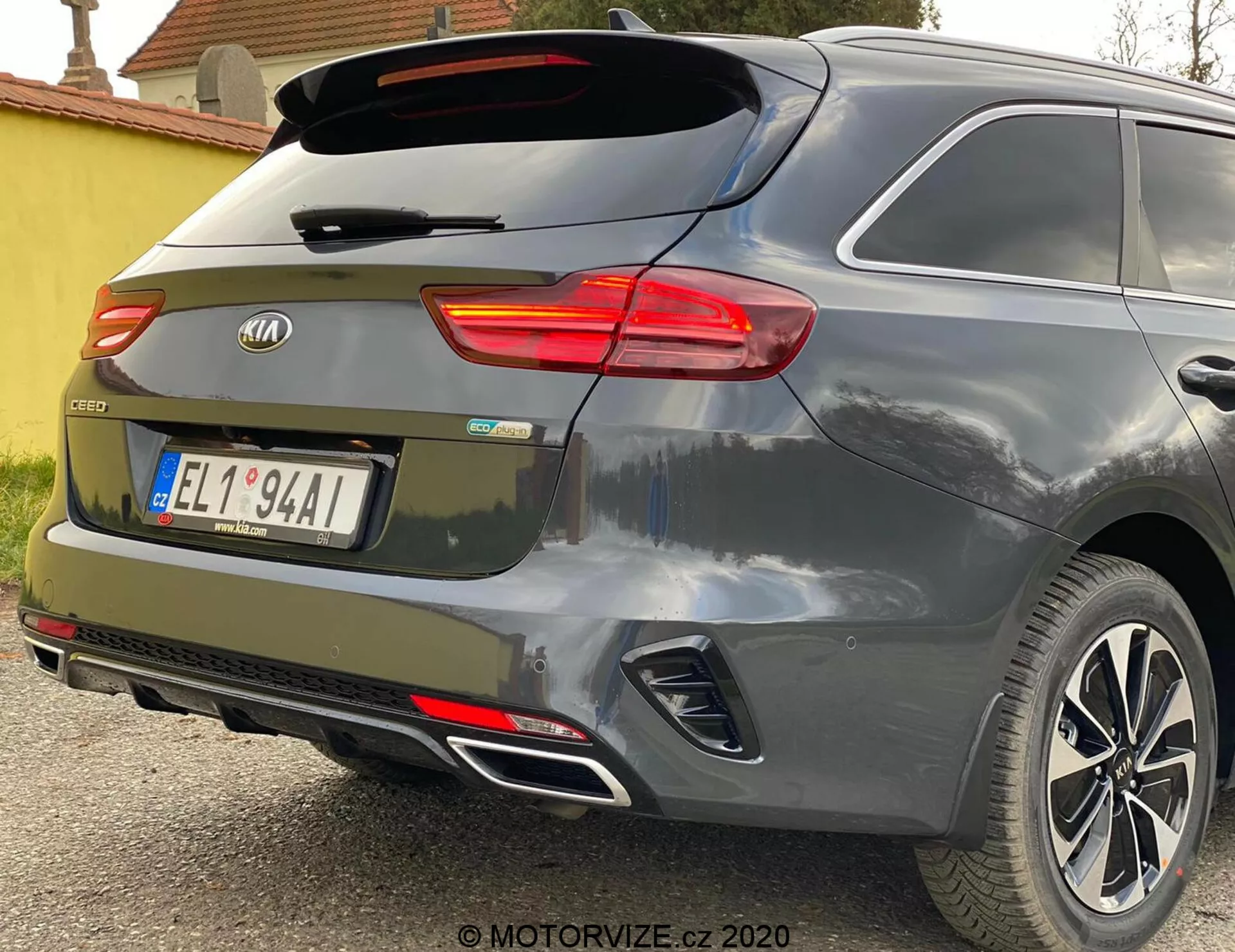 Delvis bakre vinkelvy av en Kia Ceed 2018, med fokus på det upplysta högra bakljuset, bakrutan med torkarblad, bakluckan med märkets emblem och modellbeteckningen 'CEED', integrerade reflekterande objekt i bakre stötfångare, parkeringssensorer, dubbelt avgasrör och lättmetallfälgar mot en metallisk grå eller silverfärg.