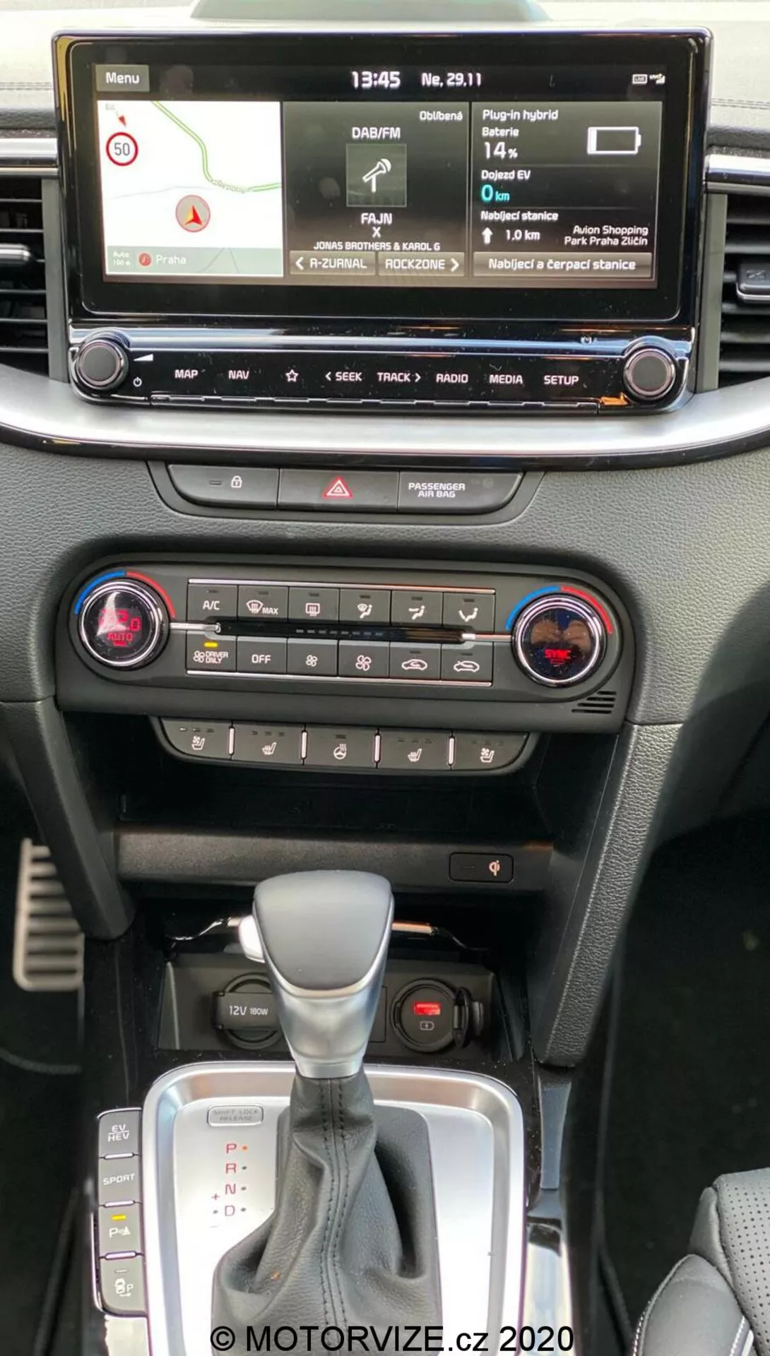 Interiör av Kia Ceed 2018 som visar den centrala konsolen från passagerarens perspektiv i främre raden, med digital infotainmentdisplay med navigeringskarta, mediespelare, fordonsinställningar, klimatkontroller med tvåzonsystem, varningsljusknapp, automatisk växelspak med läderskydd och plats för nyckelfri startknapp.