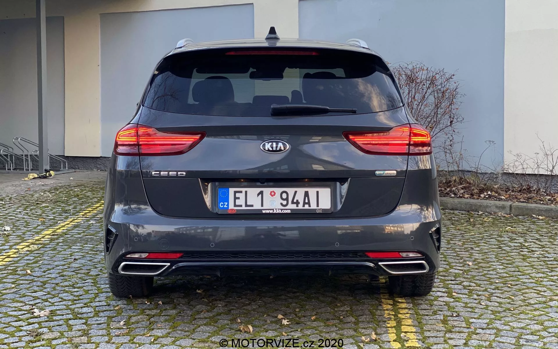Bakifrån vy av en Kia Ceed 2018 parkerad på en kullerstensyta nära en trottoarkant. Bilen, fotograferad på dagen från en rak vinkel i ögonhöjd, visar upp sina delade LED-bakljus, bakruta med torkare, tillverkarens emblem, modellnamn, registreringsskylt, bakre stötfångare med reflexer och parkeringssensorer, dubbla avgasrör och mörkgrå metalliclack. I bakgrunden syns en byggnad, vilket antyder ett bostads- eller kommersiellt område.