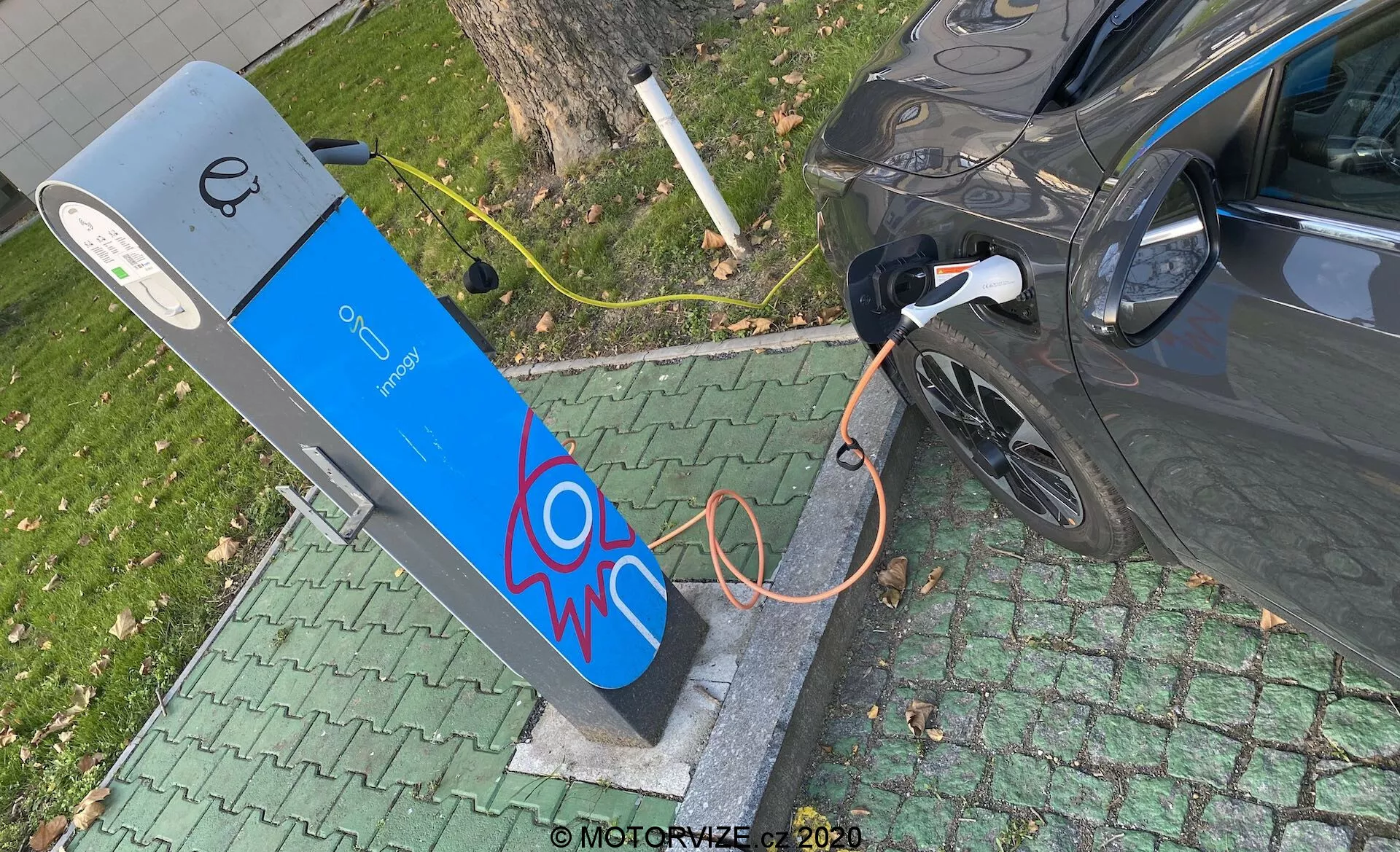 Högvinkelvy av ett modern elfordon som laddas vid en offentlig laddstation. Bilden fångar den högra bakre delen av bilen, med fokus på laddningsporten med den anslutna tjocka orange kabeln. Laddstationen har en blå fasad med en grafisk avbildning av en elektrisk gnista och en skärm högst upp, vilket tyder på användargränssnittselement. Scenen är placerad i en grön parkliknande miljö, vilket tyder på en bekväm stads- eller förortslokalisering.