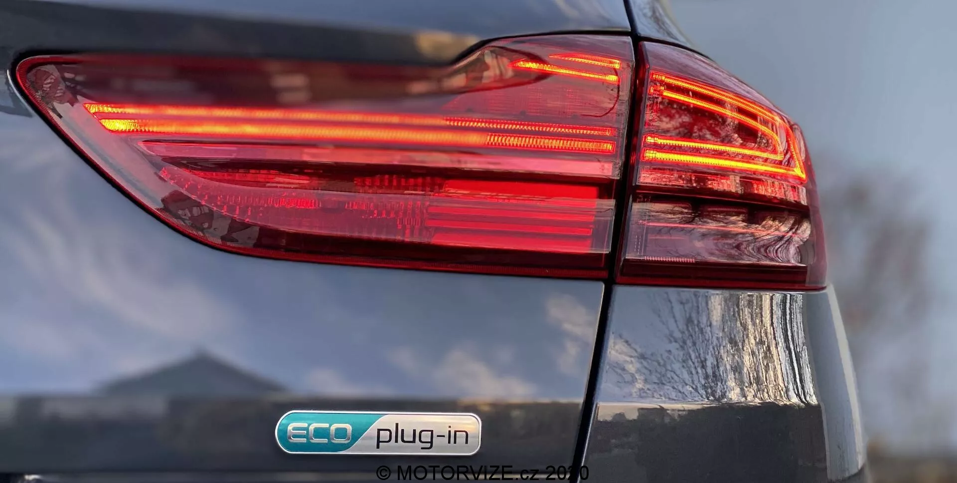 Närbild på Kia Ceed 2018s bakre vänstra sida, med fokus på det upplysta bakljusklustret med en flerskiktsdesign. 'ECO plug-in' märket indikerar att det är ett laddhybridfordon. Den glansiga finishen och delvis synliga bagageluckan lägger till detaljer till bilens övergripande design.