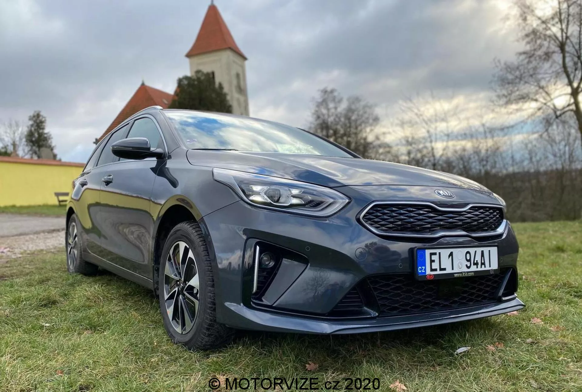 Detaljerad vy av en modern Kia Ceed 2018 hatchback fotograferad från låg vinkel i tre fjärdedelar framifrån, som visar en framträdande grill, vinklade LED-strålkastare och sidoprofilen upp till förardörren. Bilen är mörk, sannolikt djupt blå eller svart, med en glans som framhäver dess välskötta skick. Anmärkningsvärda designfunktioner inkluderar ett mönstrat frontgaller, skarpa strålkastare med klara linser, uttalade aerodynamiska element på den nedre främre stötfångaren, fler ekrade lättmetallfälgar, sidospeglar i karossfärg med integrerade blinkers och en sportig sidokjol. Bilens hållning antyder en balans mellan komfort och sportighet. Naturligt dagsljus med mjuk belysning förstärker detaljerna och i bakgrunden finns ett naturlandskap med en kyrka eller ett torn synligt.