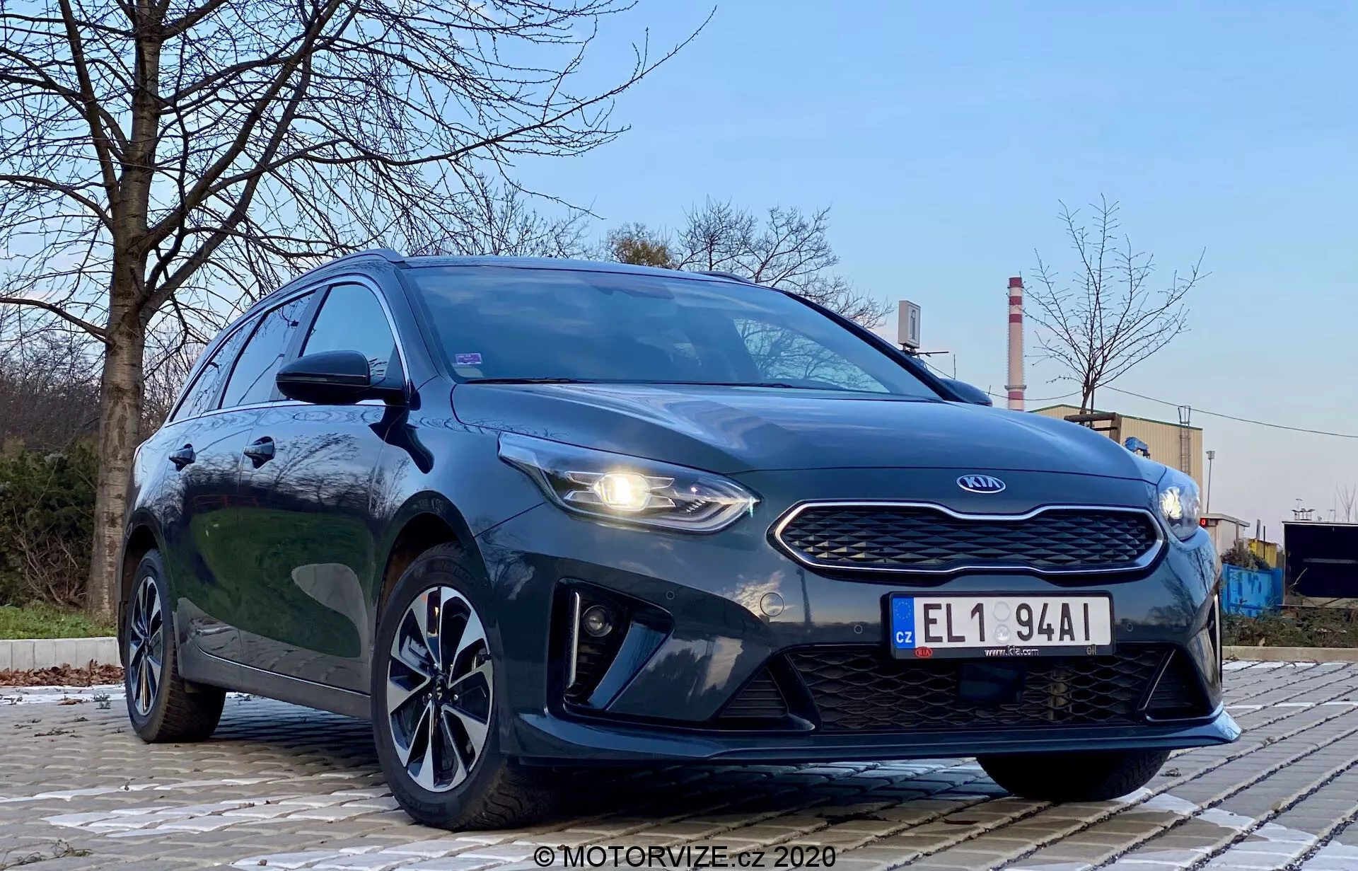 Fotografi av Kia Ceed 2018 tagen från en låg, framåt-vänster vinkel som betonar fordonets grill, strålkastare och främre fasadens design. Bilens aggressiva stil är framhävd av framträdande strålkastare, en tiger-nose grill och huvens konturer. Bilden visar även LED-dagljus, den aerodynamiska främre stötfångaren och stilen på lättmetallfälgarna. Utemiljön med träd och industriella strukturer i bakgrunden ger kontrast. Fordonets mörka, metalliska färg är glansig och väl upplyst, med reflektioner som indikerar en ren yta. Bilden fokuserar på bilen med ett dramatiskt lågvinkelperspektiv under mjuk utomhusbelysning.