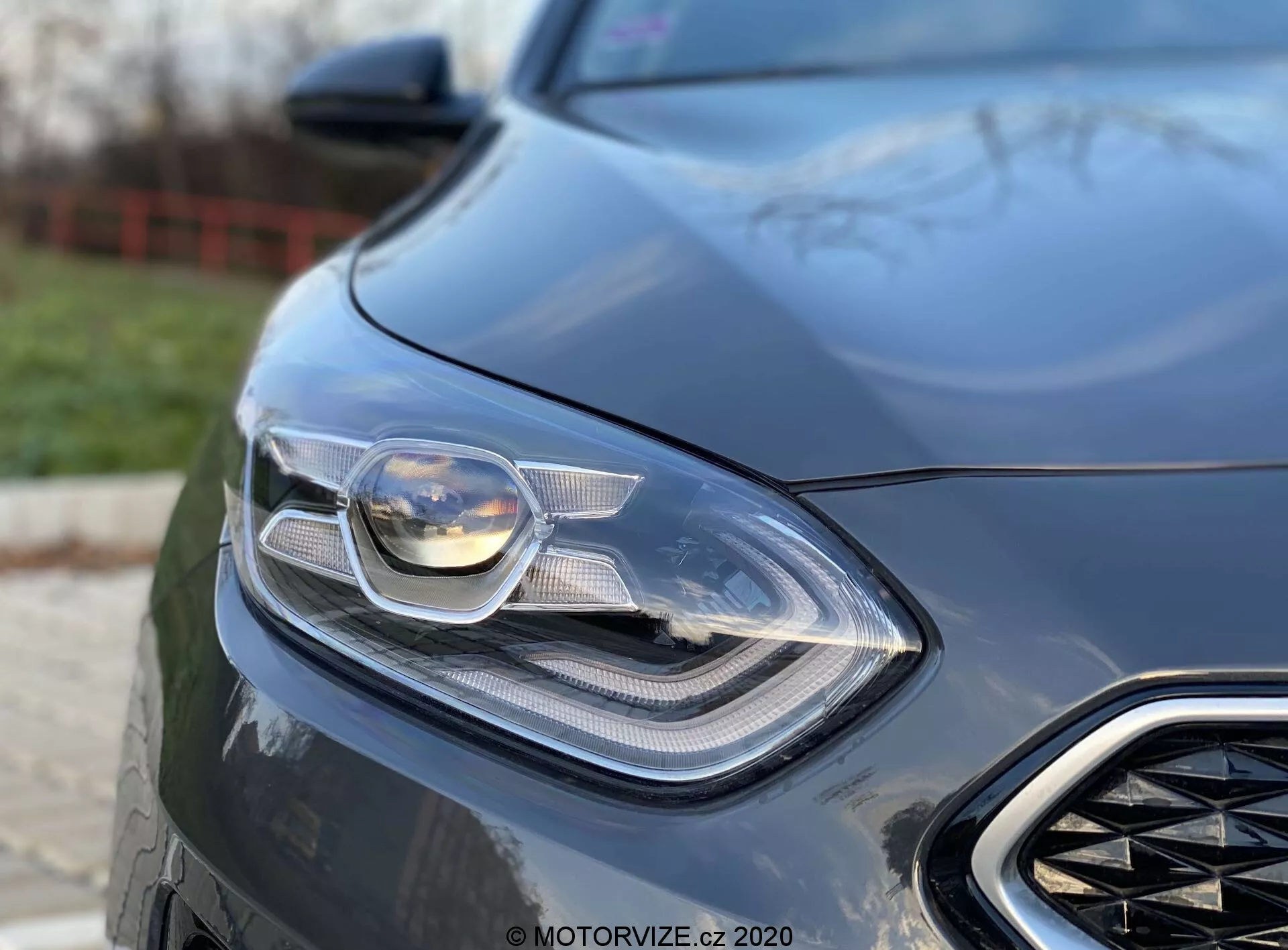 Detaljerad närbild av den vänstra frontdelen av en Kia Ceed 2018, med fokus på dess sofistikerade strålkastare och en del av grillen. Bilden är tagen från en låg vinkel, vilket framhäver bilens eleganta och moderna design med synlig avancerad belysningsteknik, möjligen inklusive LED- och adaptiva strålkastare. Den suddiga bakgrunden accentuerar bilens aerodynamiska frampartielement och lyxkänsla, vilket tyder på en prestandainriktad bil.