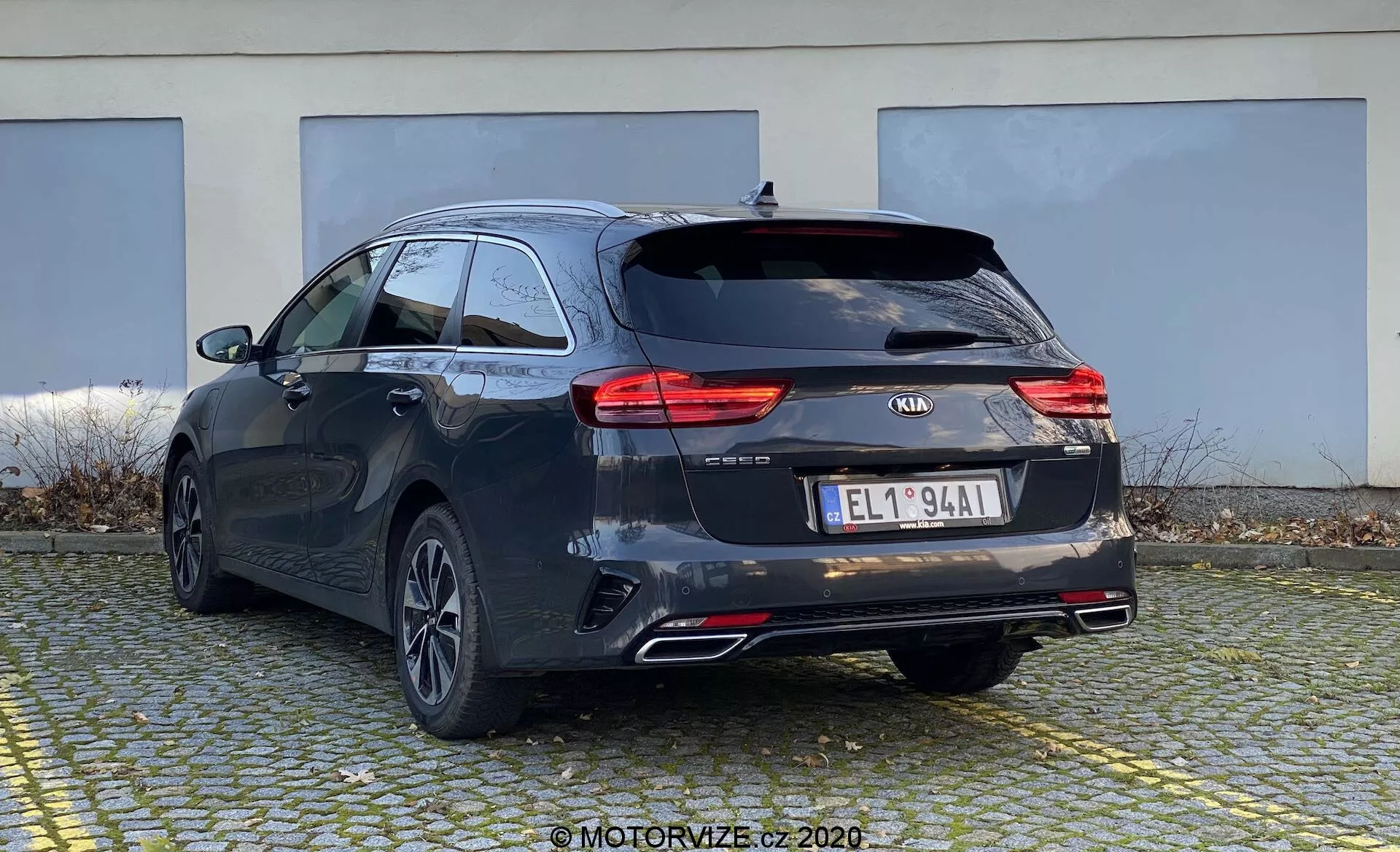 Bilden visar en Kia Ceed 2018 från en trekvart bakre vinkel, som framhäver både baksidan och sidan av kombibilen. Bilen har en mörk nyans, möjligen svart eller mörkgrå, med distinkta karaktärslinjer och ett något sluttande tak för en sportig estetik. Synliga är moderna LED-bakljus, Kia-logotypen, modellnamnet 'CEED', ytterligare märkning, en europeisk stil registreringsskylt, bakre fälgar med många ekrar, dubbla avgasrör och en bakre torkare. Troligen finns en backkamera och parkeringssensorer som inte är synliga. Liten takspoiler och takräcken indikerar extra lastalternativ. Bilen är parkerad på ett asfalterat område med en byggnad i bakgrunden, som visar bak- och delvis sidoprofilen från en något låg vinkel.
