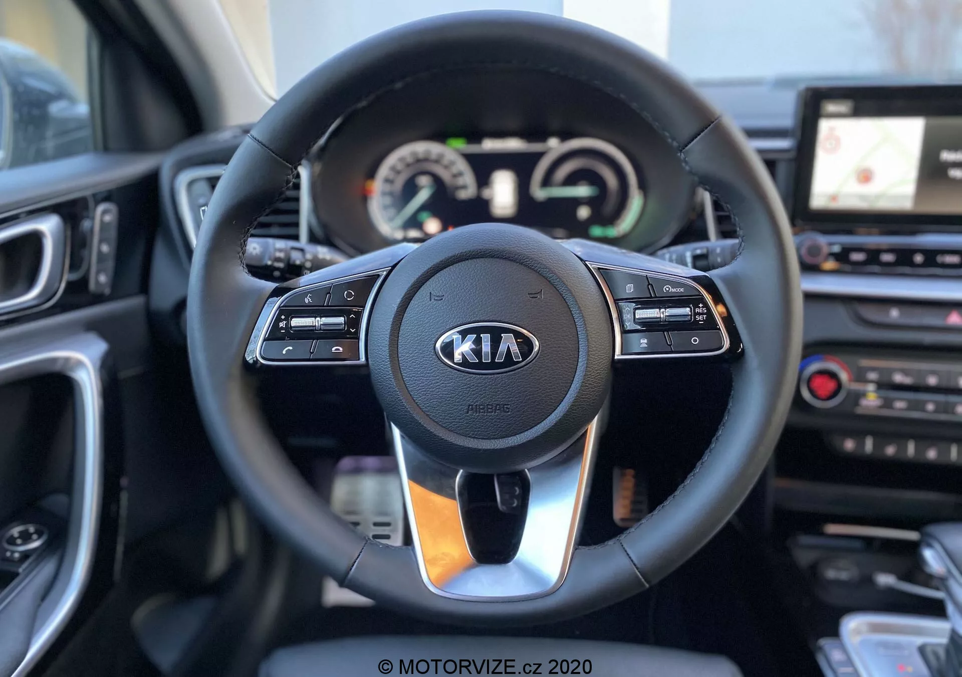 Interiörbild av en Kia Ceed från 2018 som fokuserar på ratten och instrumentpanelen från förarens perspektiv. Den moderna ratten har monterade kontroller för farthållare, multimedia interaktion och handsfree-kommunikation, med Kia-logotypen tydligt synlig. Bakom ratten finns en instrumentpanel med både analoga mätare och en digital display som visar hastighetsmätare och varvräknare, tillsammans med en multifunktionell display. Till höger om ratten syns en del av mittkonsolen med ett pekskärmsbaserat infotainmentsystem och GPS-navigation, samt kontroller för klimatanläggningen. Bilden är klar och skarp, med ett grunt skärpedjup som framhäver ratten och dess omedelbara omgivning.