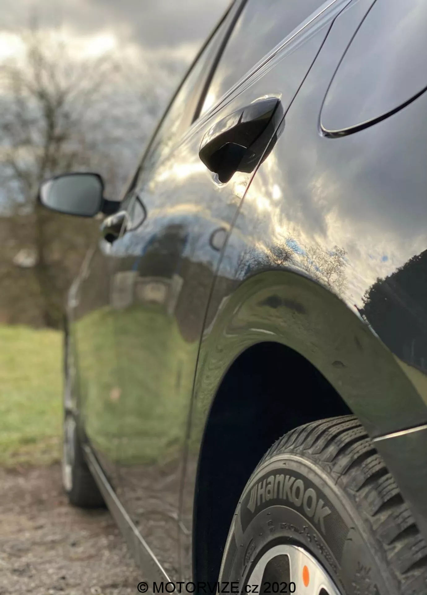 Närbild från sidan av en Kia Ceed 2018, som framhäver funktioner som bakhjulet med 'Hankook'-däck, strömlinjeformat dörrhandtag och delvis suddig sidospegel. Fotot är taget från en låg vinkel med ett kort skärpedjup som skarpt fokuserar på bakdäcket och den nedre delen av bildörren, medan bakgrunden är konstnärligt suddig. Bilen har en mörk, glansig finish som speglar himlen och omgivningen, vilket visar på eleganta designlinjer och välskött kaross.