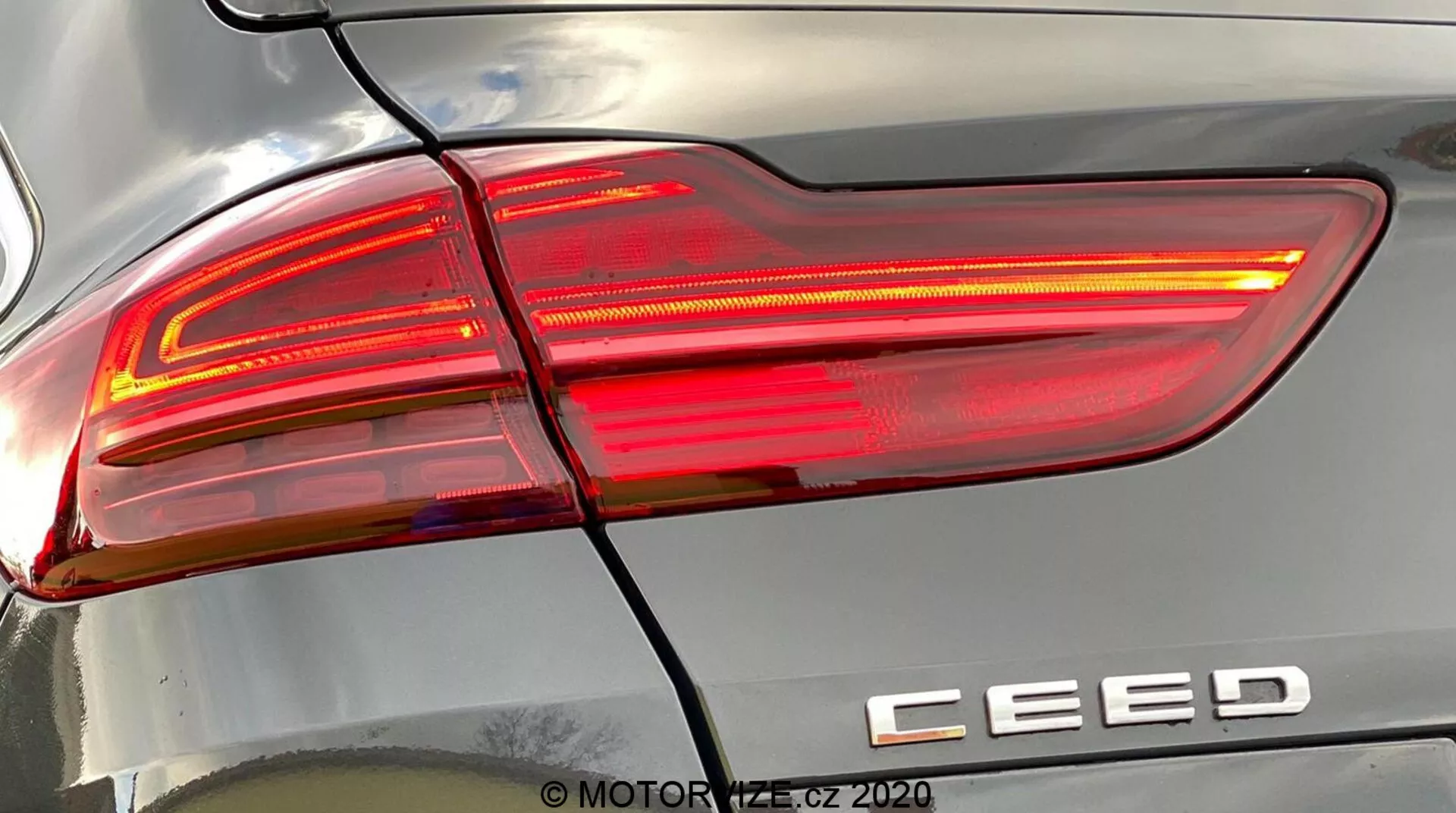Närbild med detaljerad vy av den högra bakre lyktan på en Kia Ceed 2018, upplyst, som visar modern design med skarpa, kantiga linjer, klara och röda linssektioner och modellmärkning 'CEED'. En horisontell linje separerar baklyktan från bakre stötfångaren. Reflektioner på karossen betonar ytkurvaturen. Vattenstämpel 'MOTOR1.cz ©2020'.