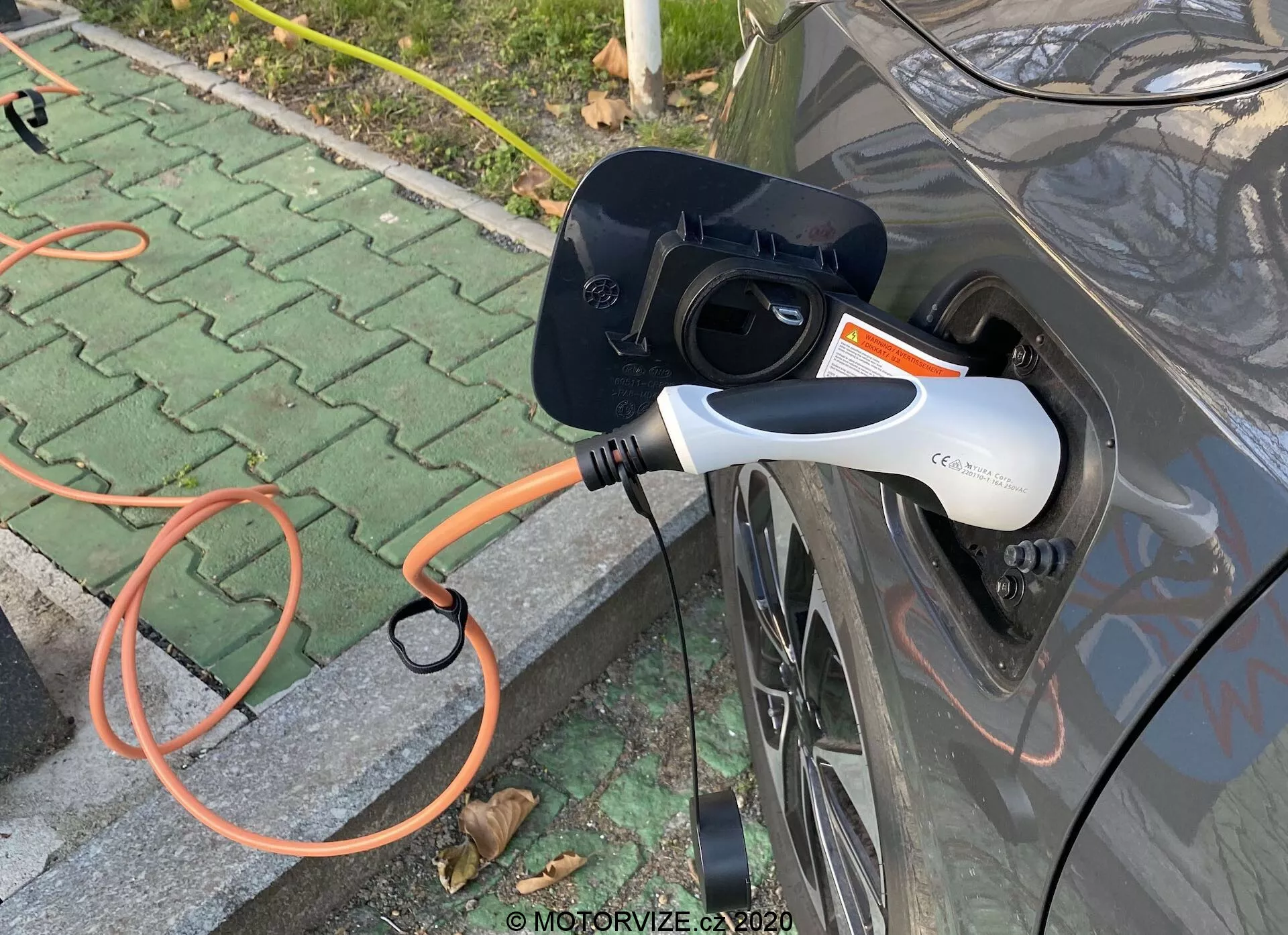 Närbild från sidan på en grå elbil (EV) som laddas, med fokus på den öppna laddporten och en orange EVSE-kontakt ansluten till bilen. Den moderna vita och svarta laddkontakten har en LED-indikatorlampa. Bilden fokuserar på laddningsinteraktionen, placerad på ett utomhusområde med några spridda löv.