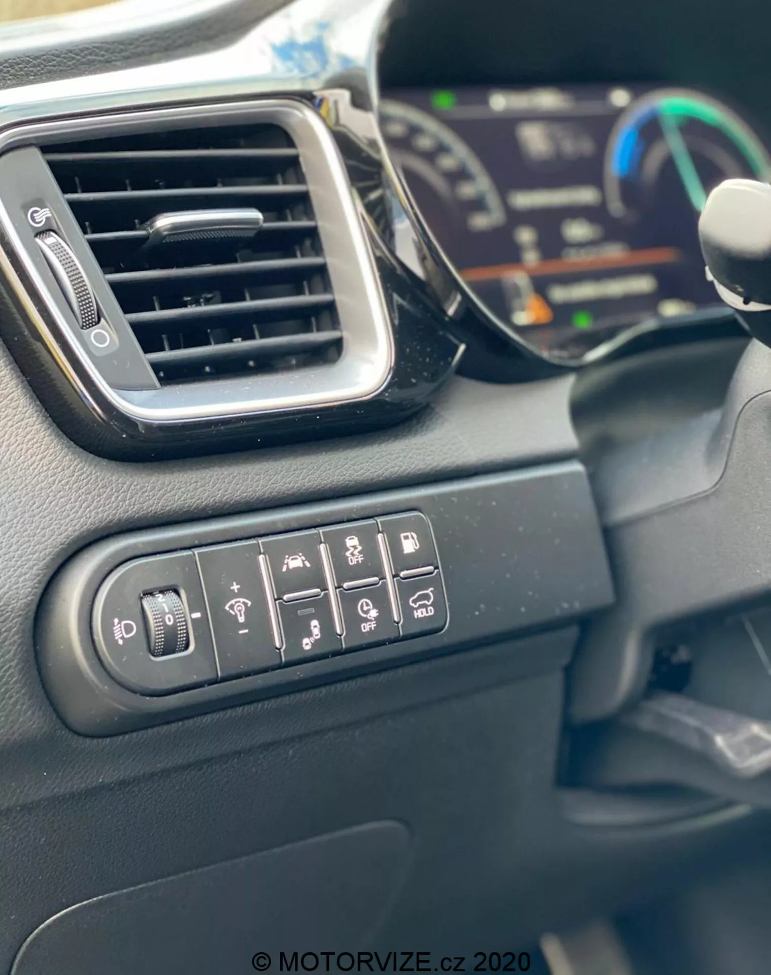 Närbild av Kia Ceed 2018 interiör som visar kontrollknappar för justering av sidospeglar, bakruteuppvärmning, rattvärme, körfältsvarning, parkeringsassistans och antispinnsystem. Bilden visar också en elegant ventilationsöppning, en del av den digitala instrumentpanelen i suddig bakgrund och kanten av ratten. Interiörmaterialen består mestadels av hårda plastmaterial med matt finish, fångade i dagsljus.