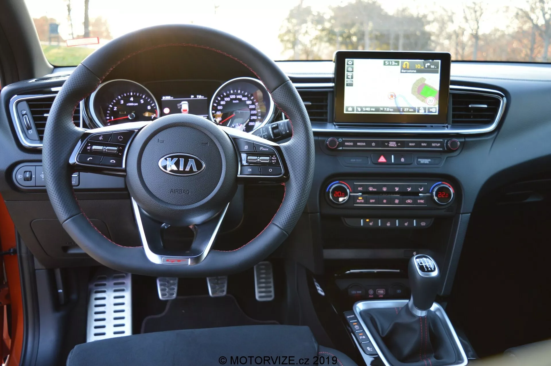 Interiörvy av en Kia Ceed 2021 från förarens perspektiv, som visar den detaljerade instrumentpanelen. Bilden framhäver tydligt ratten med Kias logotyp och monterade kontroller för ljud, samtal och farthållare. Bakom kan du se instrumentbrädan med kromkantade analoga mätare och en digital display. Den centrala infotainmentskärmen visar GPS-navigering, medan klimatkontrollknappar och vred finns under. Mittkonsolen visar växelspaken, mugghållare och en USB-port för enhetsanslutning, som framhäver modern bilteknologi.