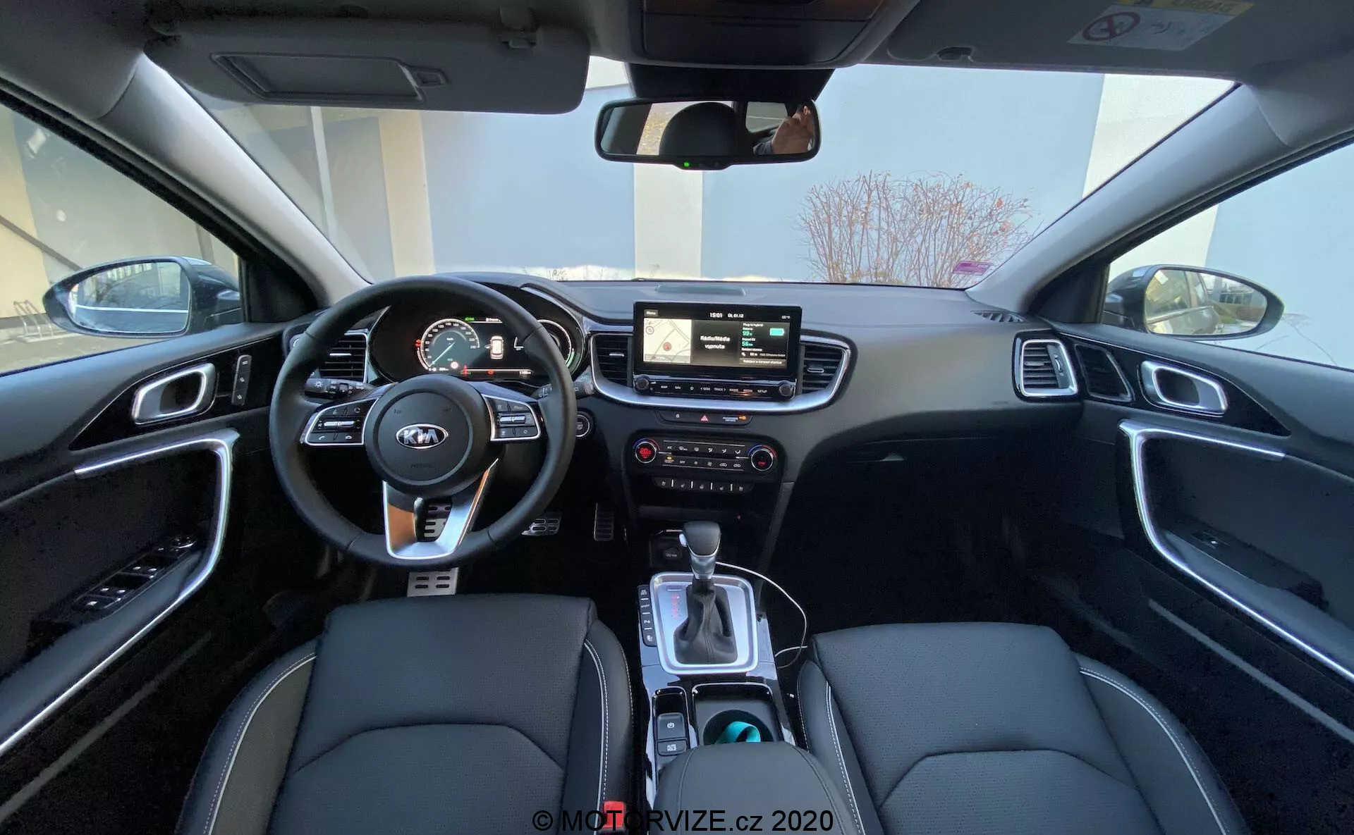 En vidvinklad bild av interiören i Kia Ceed 2018 från baksätet som visar förarområdet inklusive ratt med monterade kontroller, instrumentkluster med digitala och analoga mätare, central infotainment-system med pekskärm, manuella kontroller för infotainment och klimat, samt mittkonsol med växelspakar, körlägesväljare, mugghållare och en svart interiör med syntetiska lädersäten med vita sömmar och borstade aluminiumdetaljer.