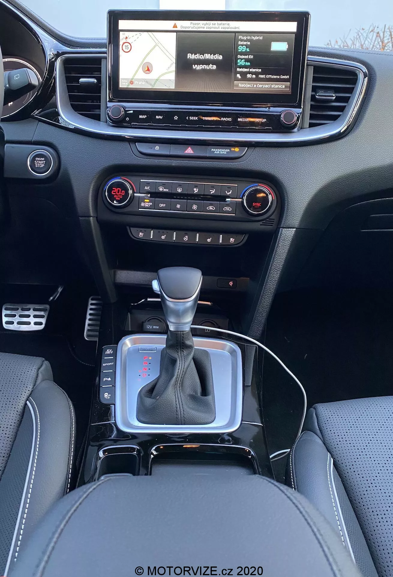 Interiörvy av en 2018 Kia Ceed från bakre sittplats med en modern instrumentpanel, pekskärms infotainmentsystem som visar en navigationskarta, knappar och reglage för ljud och klimatkontroll, centralt ventilationsgaller, växelspak i neutralt läge med silverfärgade detaljer och svart läderomslag, litet förvaringsutrymme nära växelspaken eventuellt för 12V-uttag eller USB-portar, lädersäten med detaljerade sömmar.