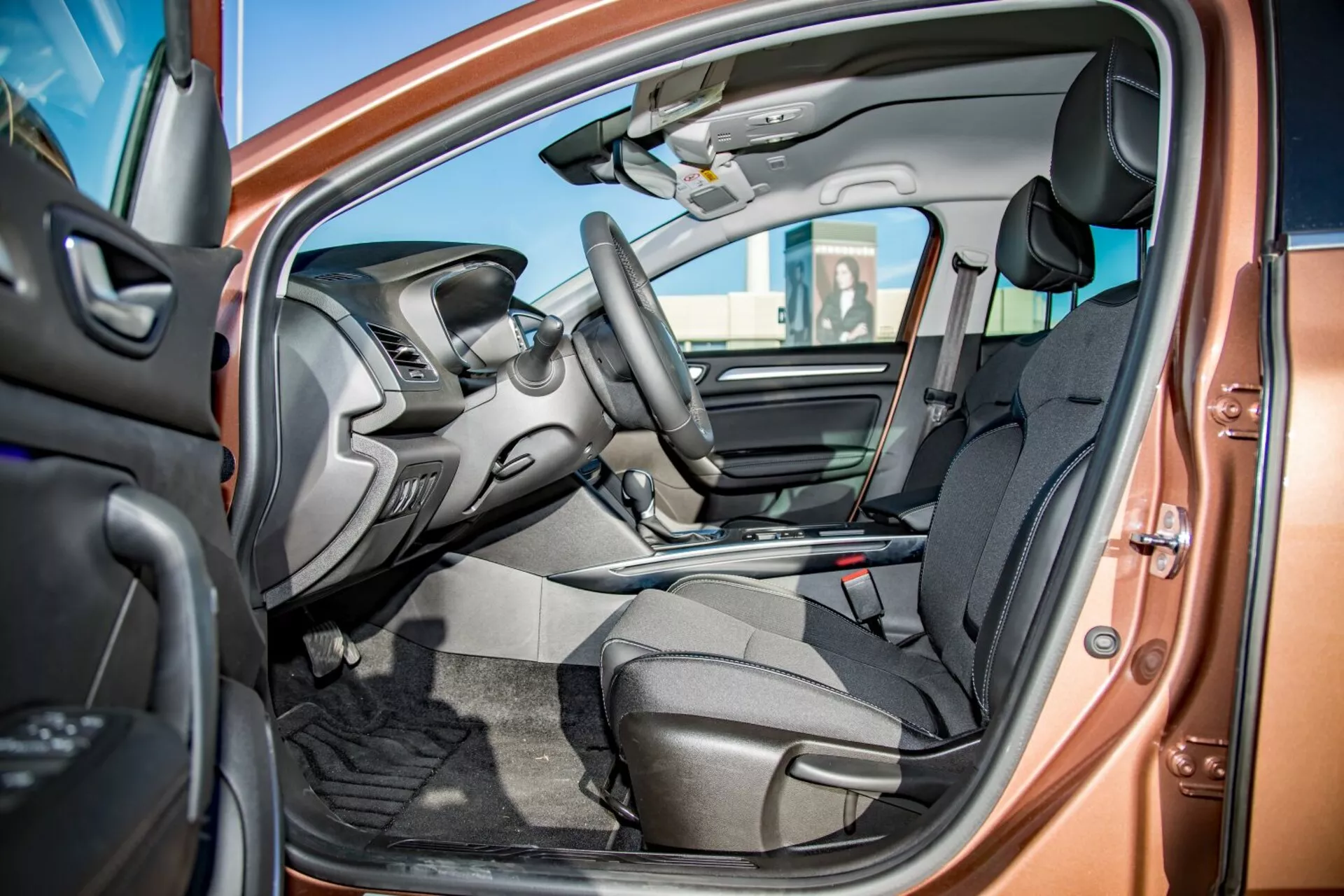 Interiörvy av Kia Ceed Pro-Cee'd 2009 genom den öppna förardörren som visar framsäten i mörkt tyg, integrerade rattkontroller, delvis synlig instrumentbräda med infotainmentskärm, mittkonsol med växelspak, bilgolvsmattor och tvåtonig färgsättning