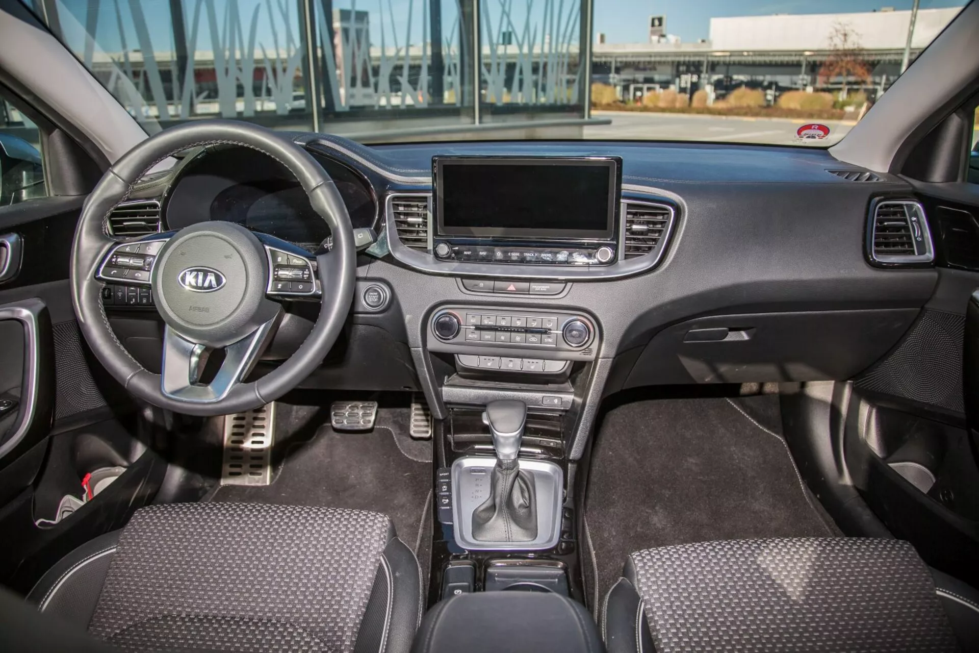 Interiörvy av Kia Cee'd från 2009 som visar hela frontkabinen inklusive multifunktionsratt med Kia-logo, instrumentkluster bakom, växelspak i parkeringsläge på mittkonsolen, klimatkontrollknappar, infotainmentskärm, tygklädda säten, och förar- och passagerarsidans dörrkontroller i naturligt dagsljus