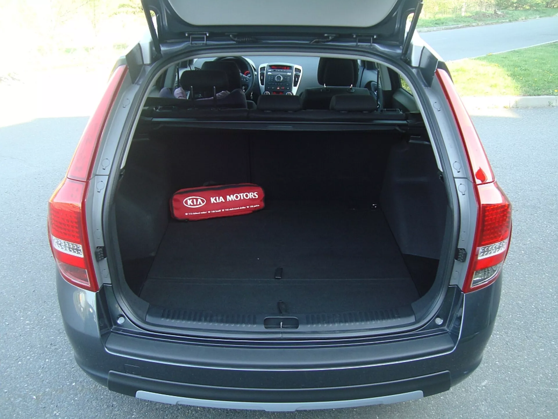 Bakre delen av bagageutrymmet på en Kia Cee'd hatchback från 2009 med öppet lock som avslöjar ett rymligt, tomt lastutrymme, hopfällbart överdrag, interiör textilmatta och Kia Motors logotyp på ett rött skyddsöverdrag, som visar fordonets design, säkerhet och teknologiska funktioner.