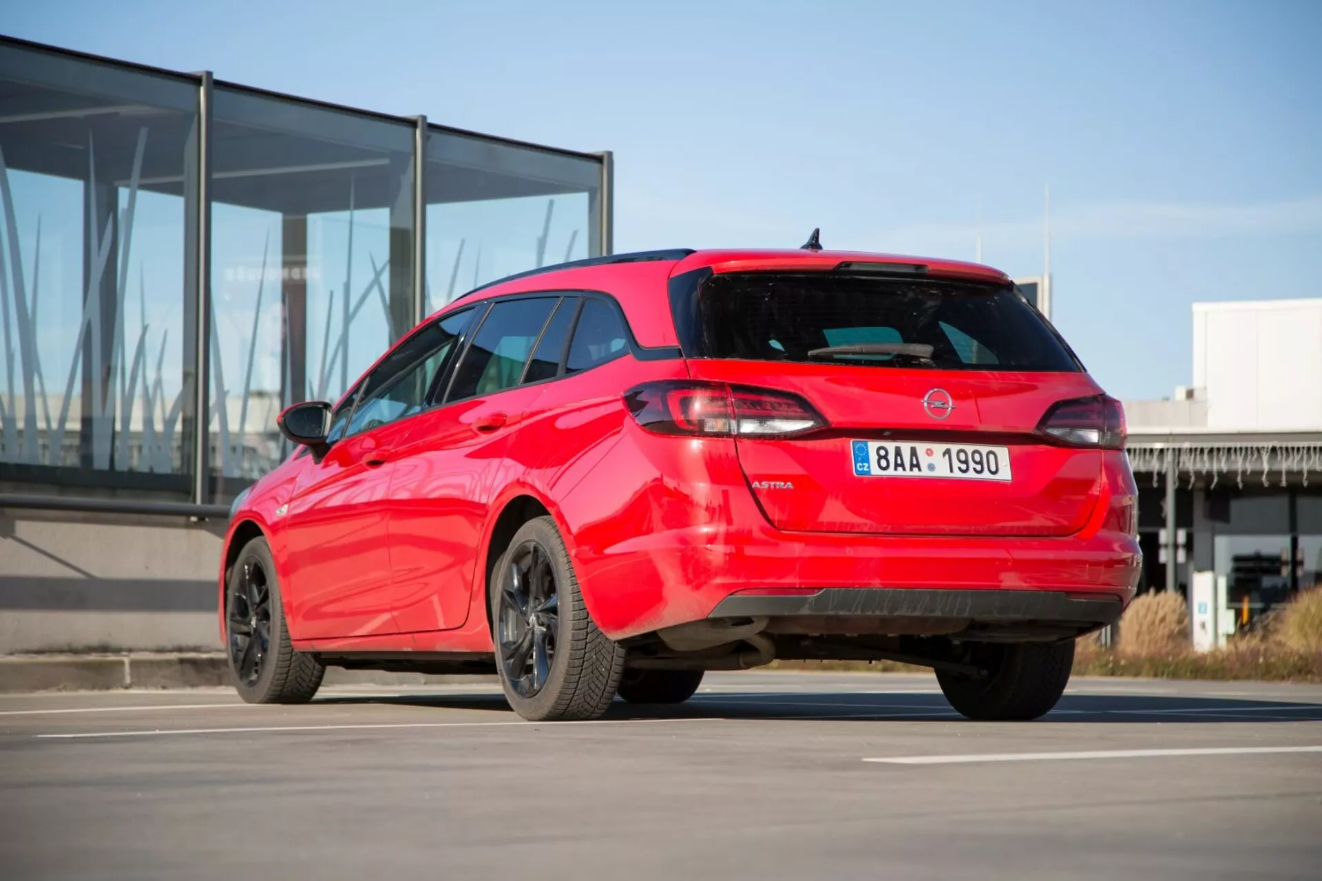 2009 Kia Ceed hatchback i rött som visar sin sportiga tre kvarts bakvy på betong med modern glasfasadbyggnad i bakgrunden, som visar distinkt bakljusdesign, takspoiler, hajfenaantenn, legeringsfälgar och möjliga parkeringssensorer.