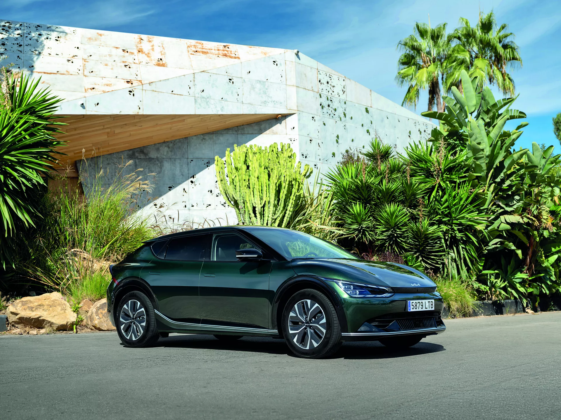 Kia EV6 2021 fångad från ett lågt vinkel vilket framhäver dess slanka design och sportiga ställning med en tredjedels fram-sidovy som visar LED-strålkastare, aerodynamisk kaross, legeringshjul, integrerade blinkers på sidbackspeglar, parkerad utomhus under ljus dagsljus med en modern byggnad och frodig grönska i bakgrunden