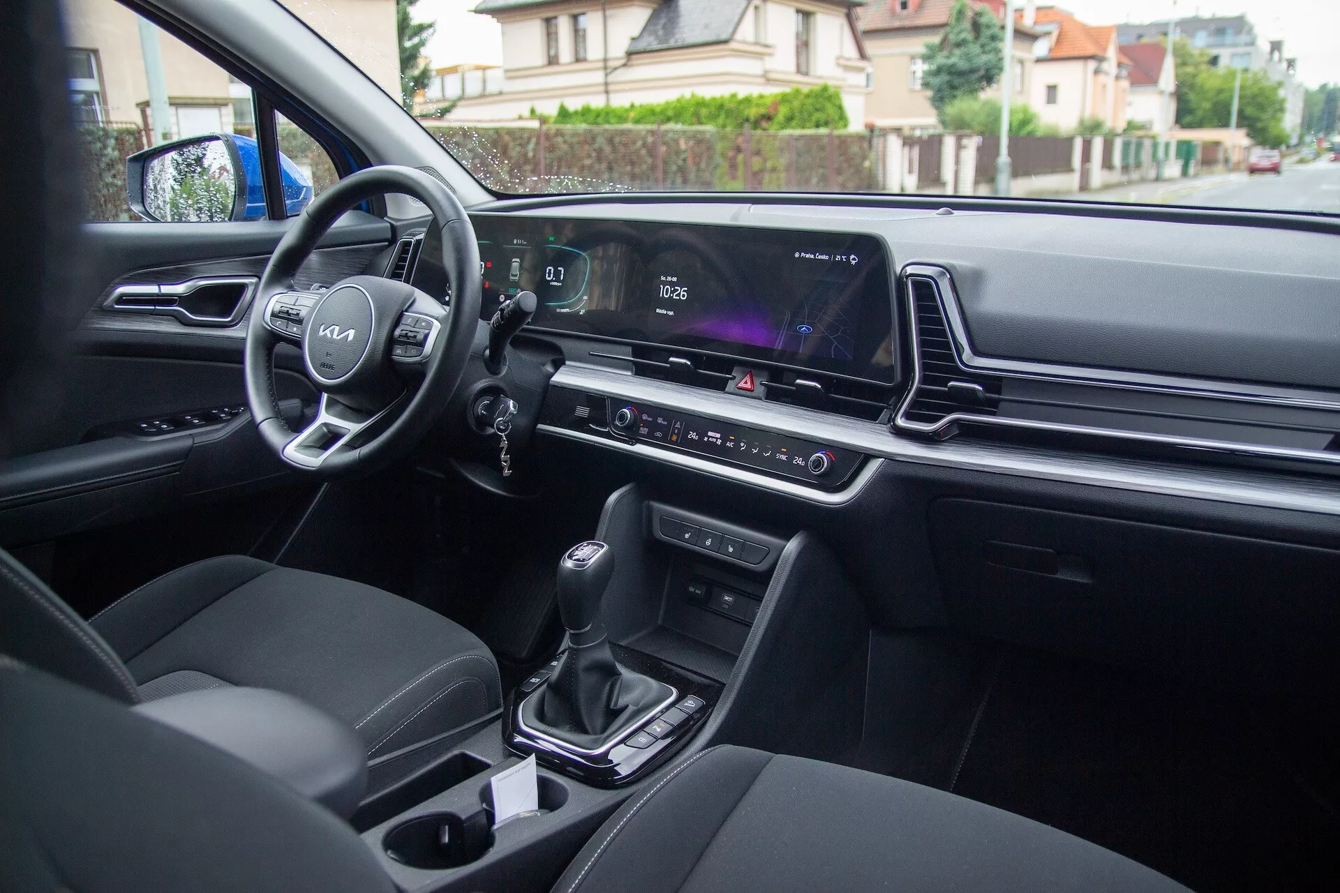 Interiörvy av Kia Sportage 2022 från baksätet som visar förardörrskontroller, ratten med logotyp och monterade reglage, digital instrumentkluster, infotainmentskärm, klimatkontroll, automatisk växelspak, mugginnehavare och moderna klädda säten