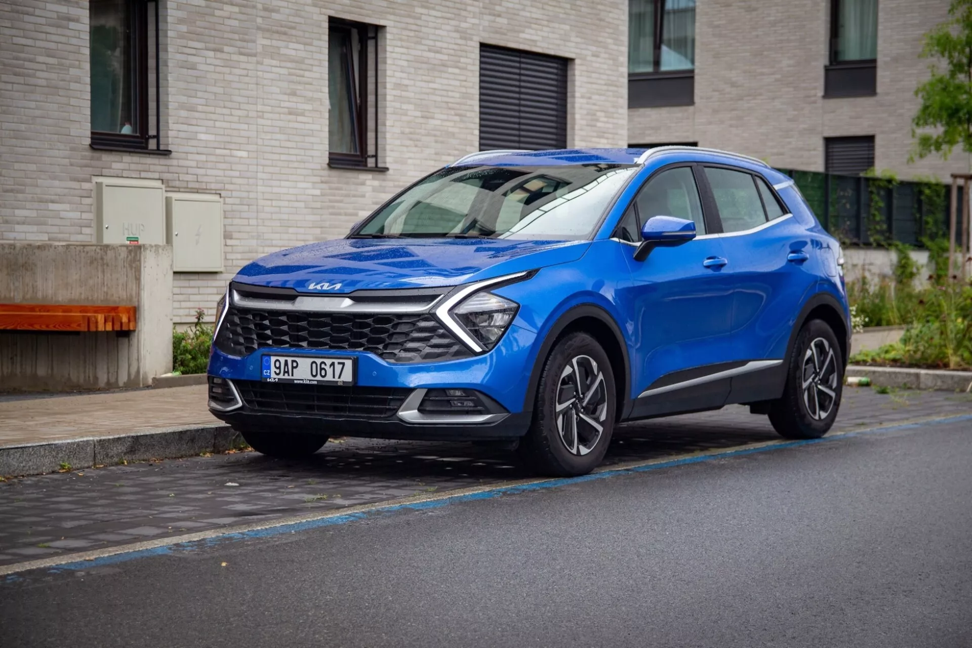 Kompakt crossover SUV Kia Sportage 2022 i blått, parkerad på en våt gata som visar sin dynamiska främre tredjedels vy med LED-dagsljus, skulpterad motorhuv och legeringshjul.