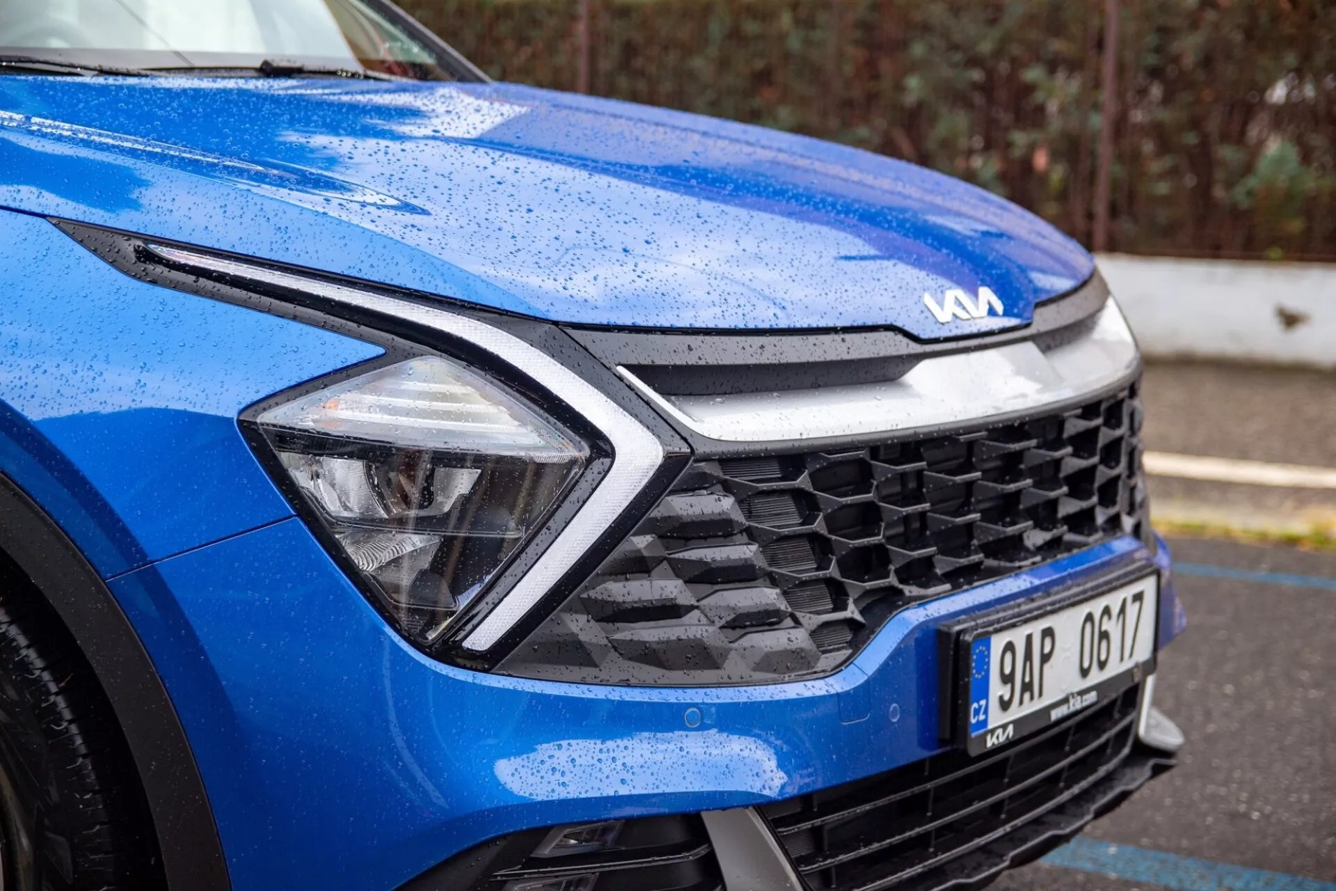 Närbild av en blå Kia Sportage 2022 fokuserad på främre vänstra kvartalet under våta förhållanden, som framhäver den dynamiska motorhuvsdesignen, LED-strålkastare, bikaketrist och avancerad sensorteknik